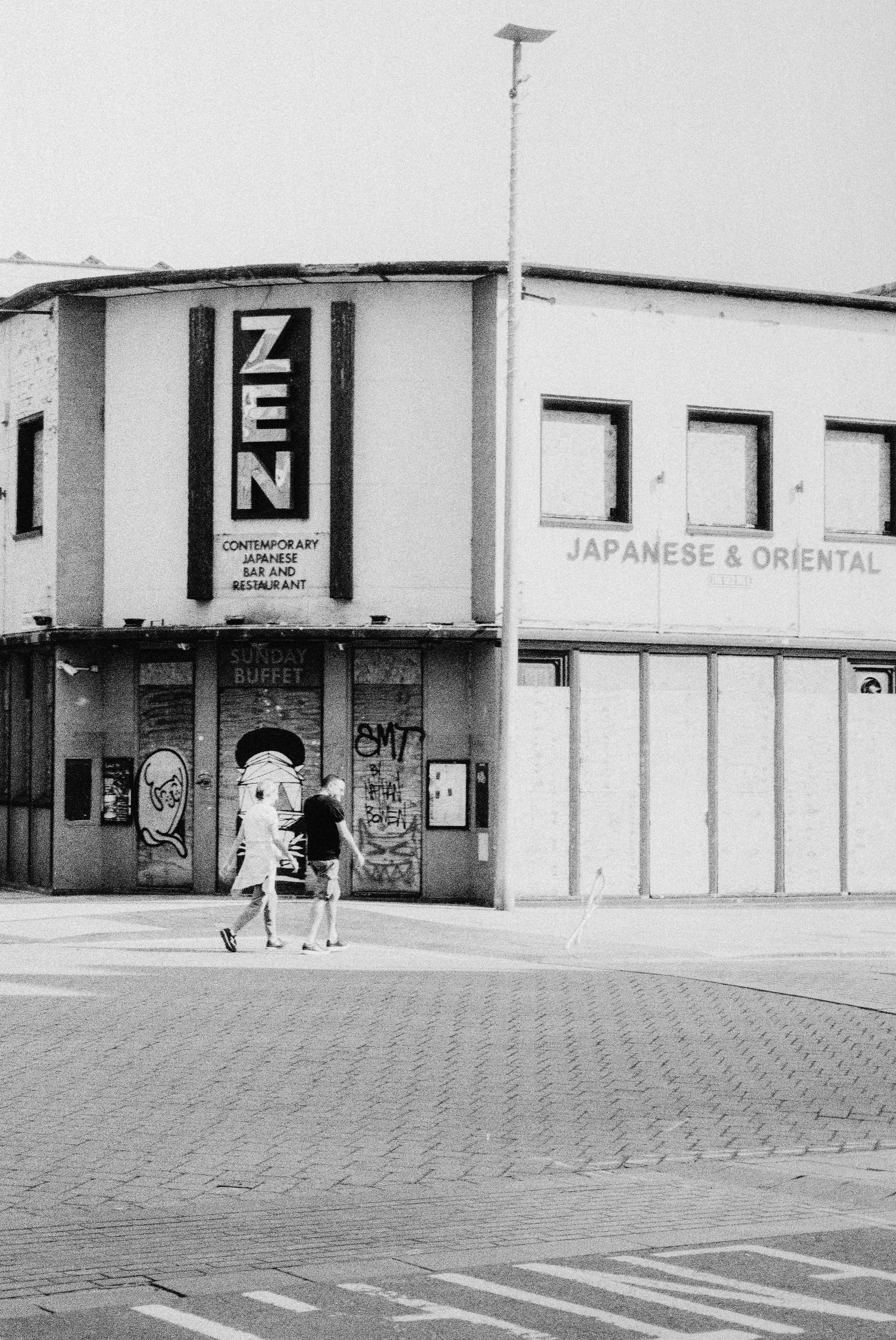 Two people walking past a restaurant with graffiti on the boarded windows