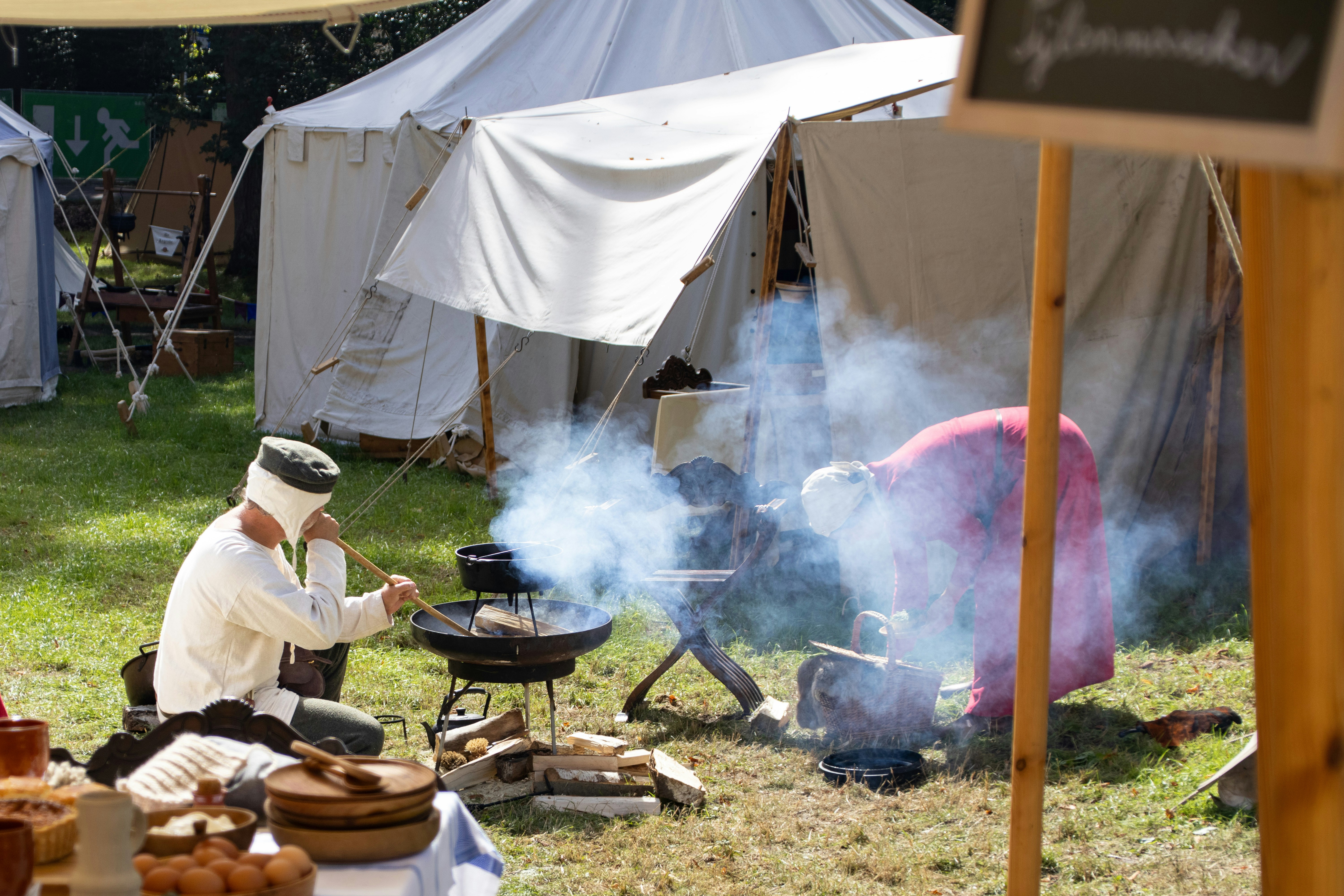 Two people in historical clothing cooking over an open fire near white canvas tents