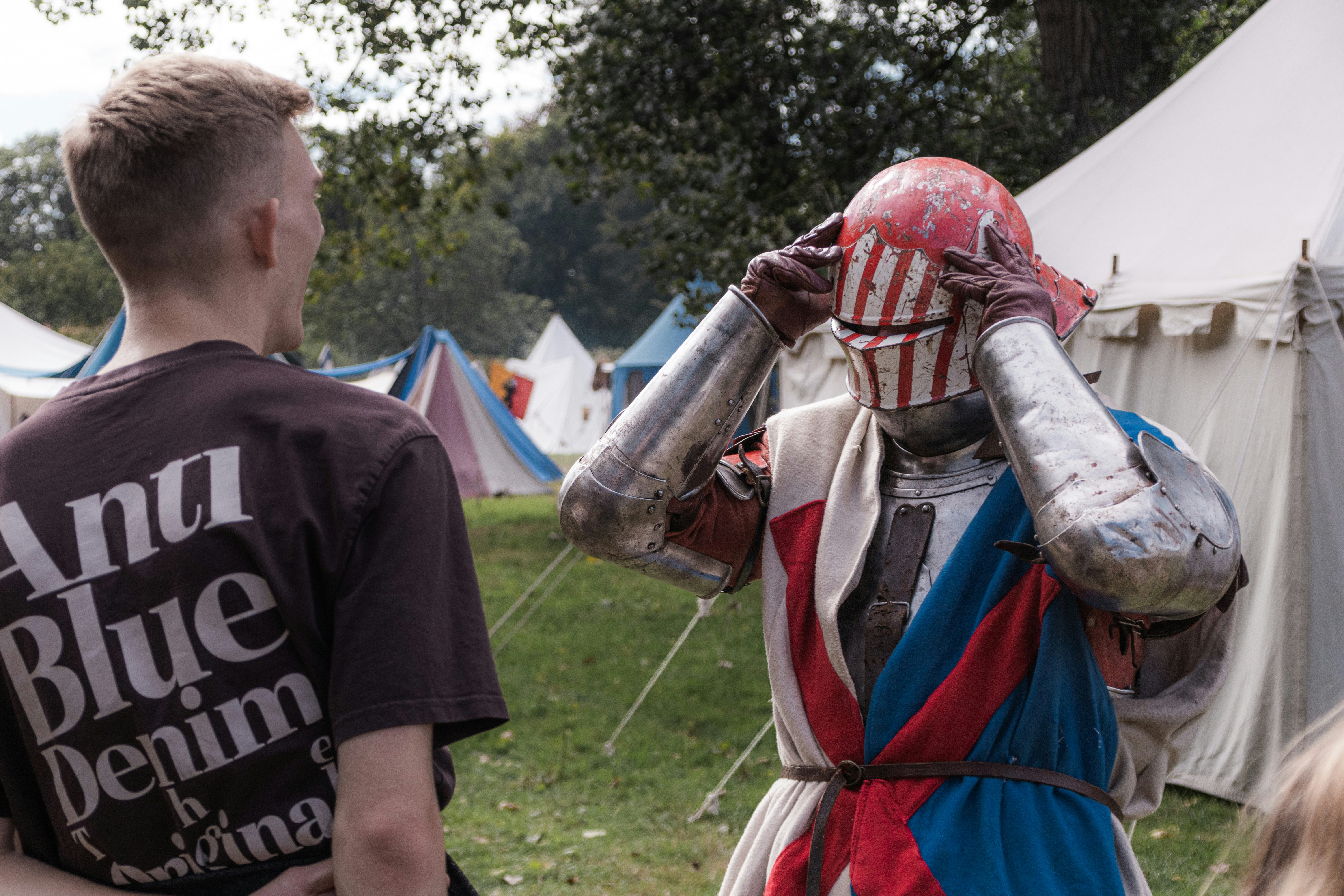A person in a brown t-shirt talking to a knight adjusting a red helmet