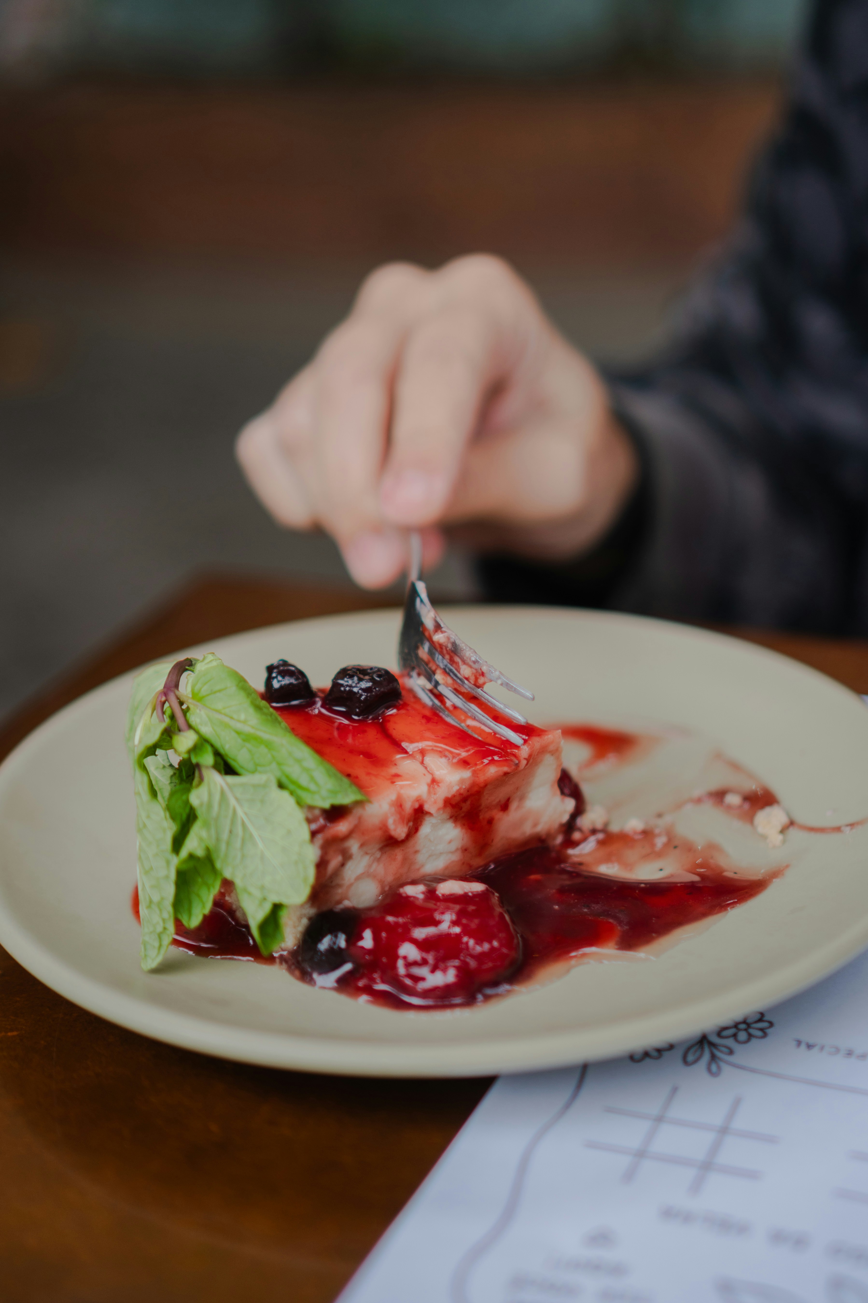 A person using a fork to eat cheesecake with berry sauce and mint