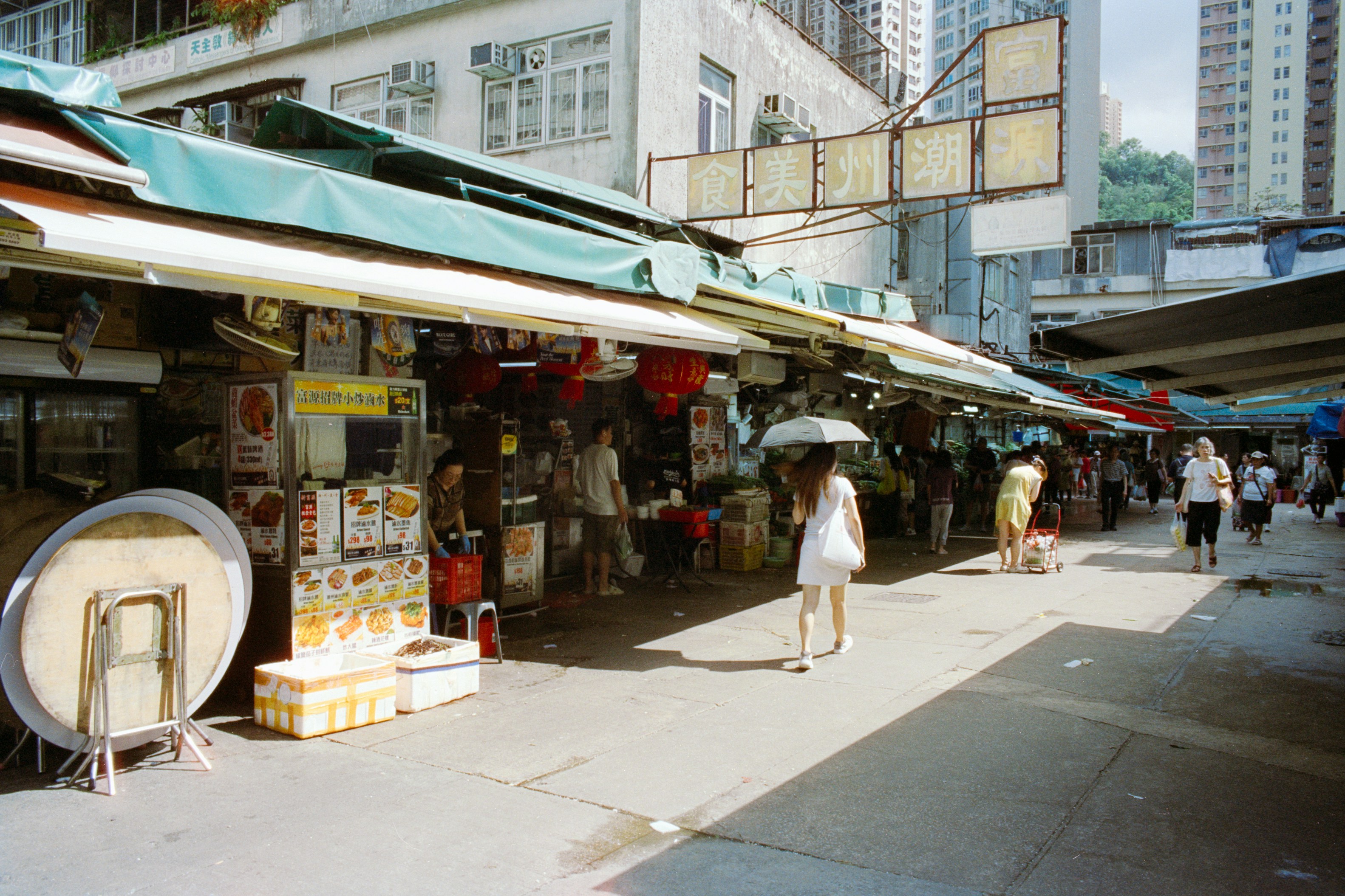 A person walking with an umbrella through a sunlit outdoor market in Hong Kong