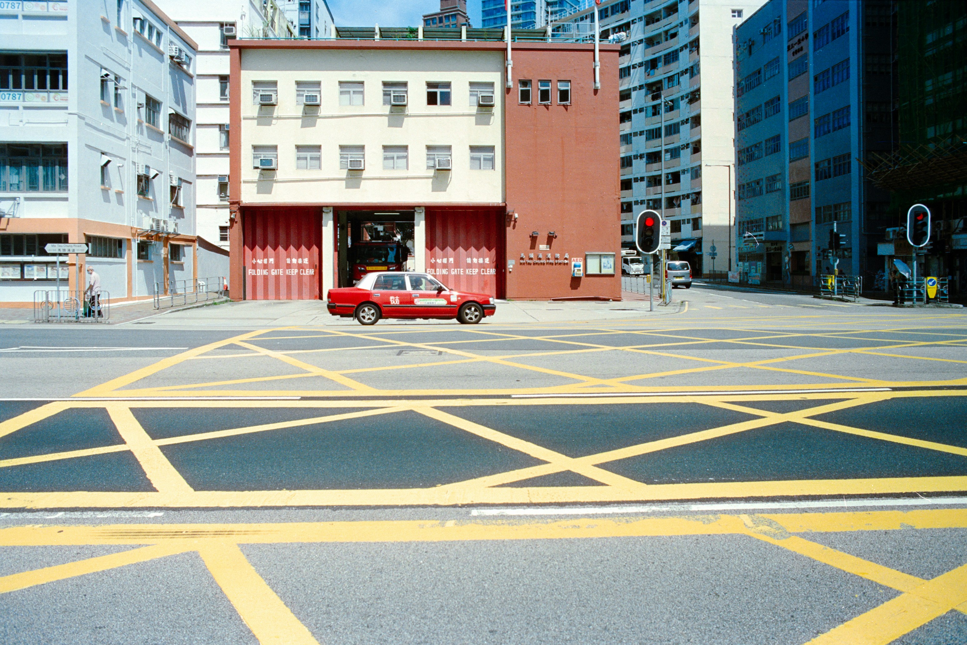 A red taxi crossing a yellow grid intersection in front of a fire station