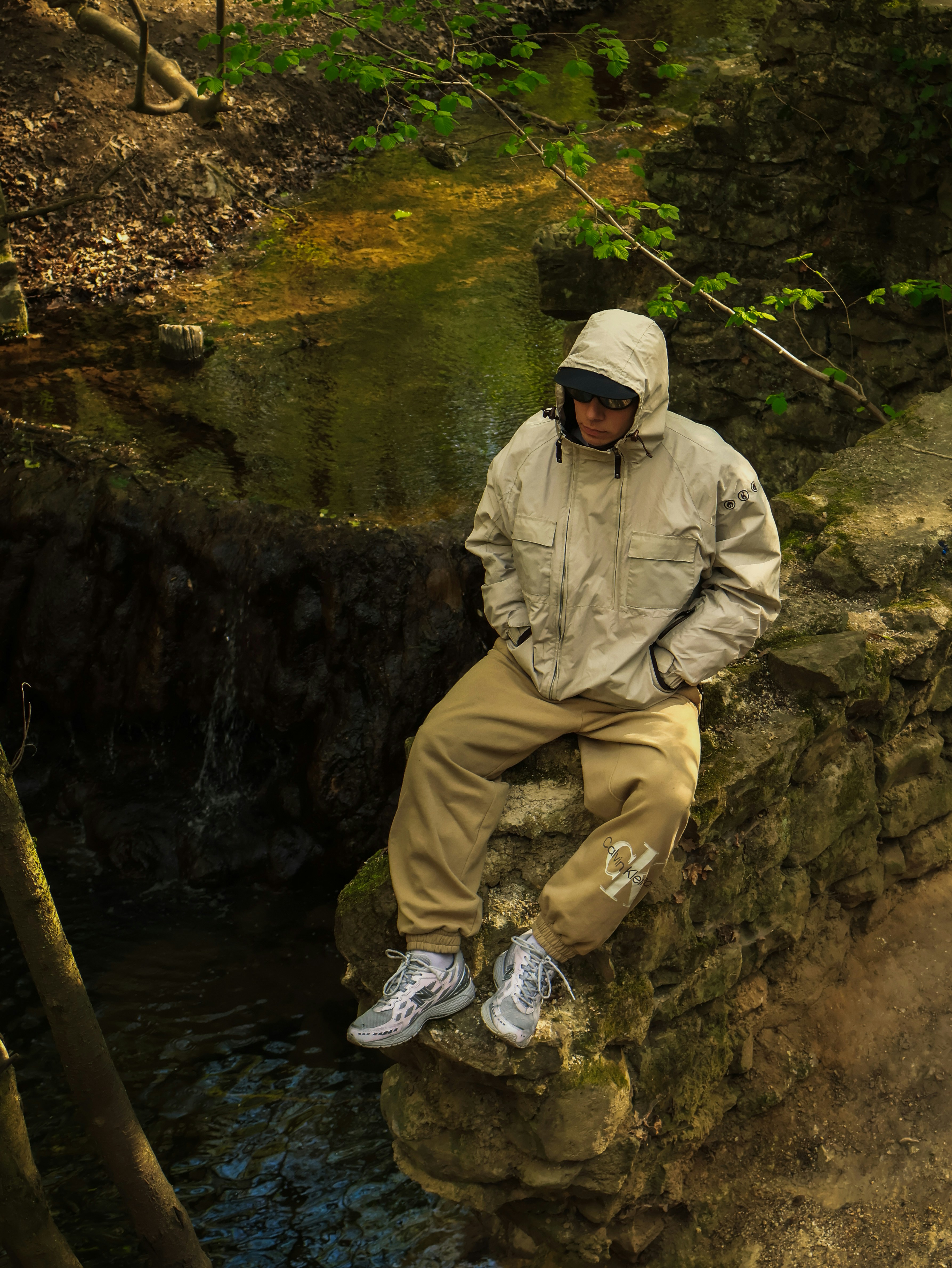A person in a beige hooded jacket and sweatpants on a mossy stone wall