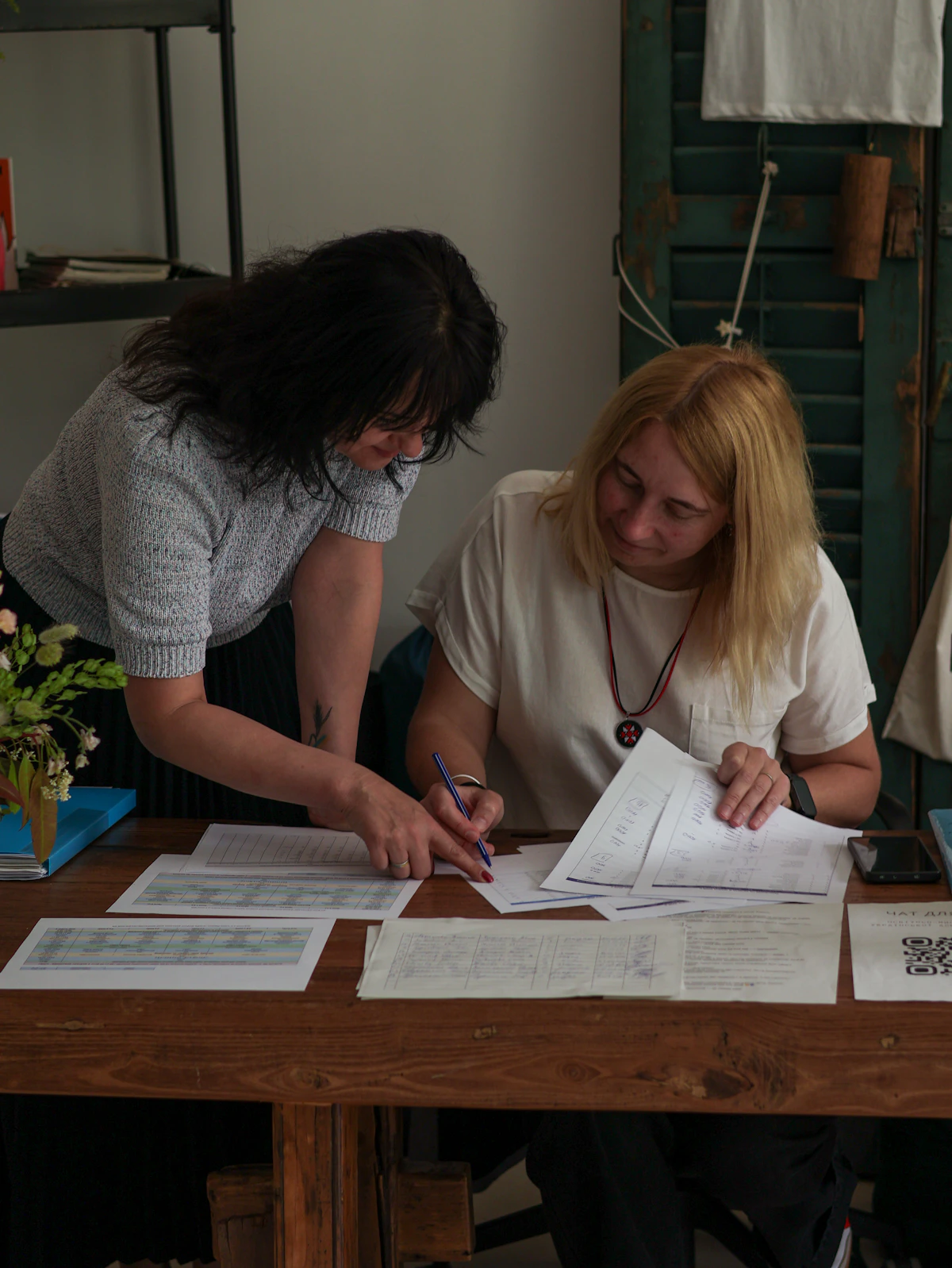 Two women at a wooden table go over printed schedules together, one leaning in to point at a sheet with her pen