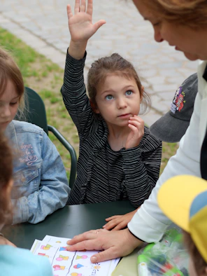 A child with her hand raised at a table with an adult and peers