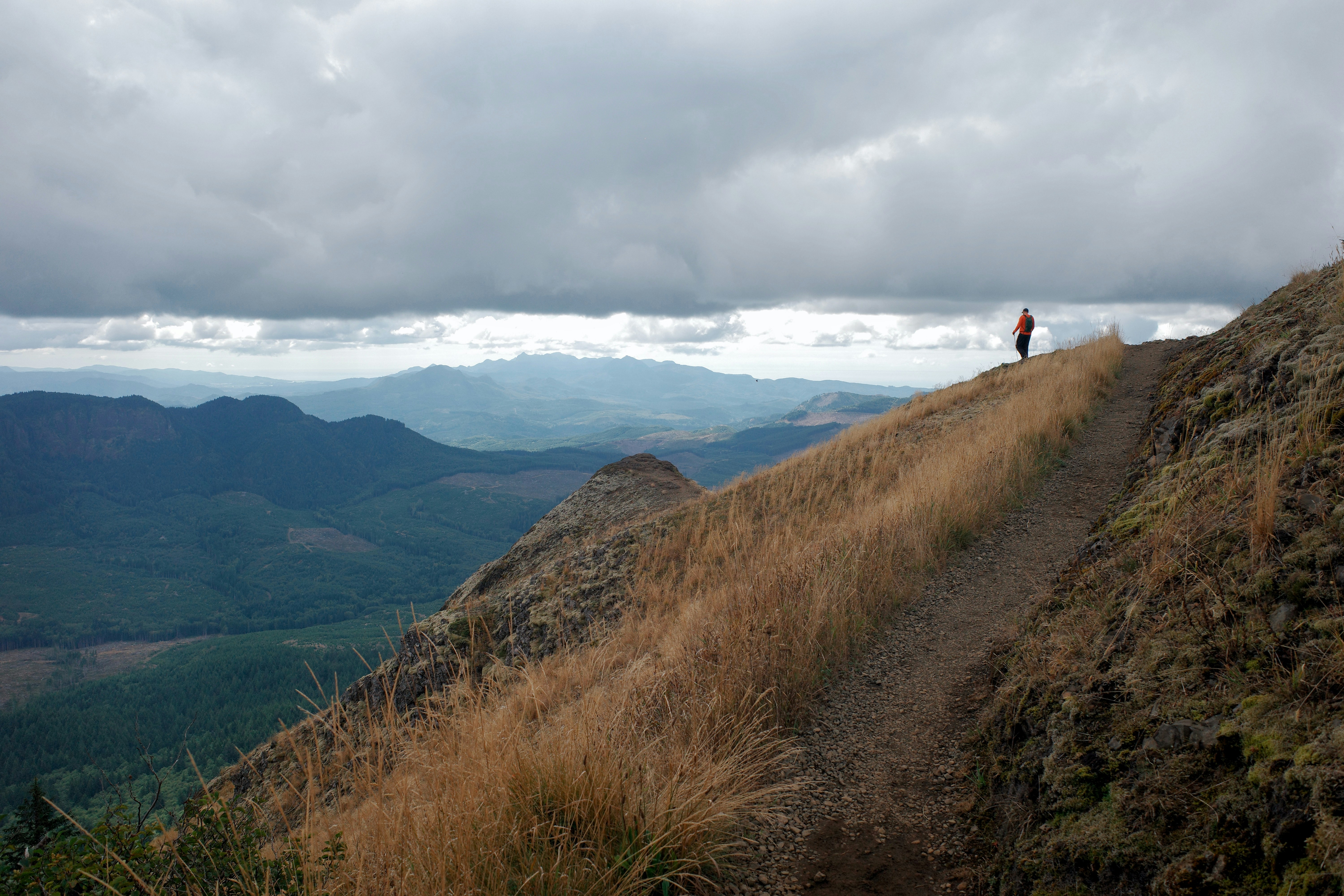 Lone hiker in an orange jacket far up a narrow dirt trail along a dry grassy ridge, under a low grey cloud ceiling
