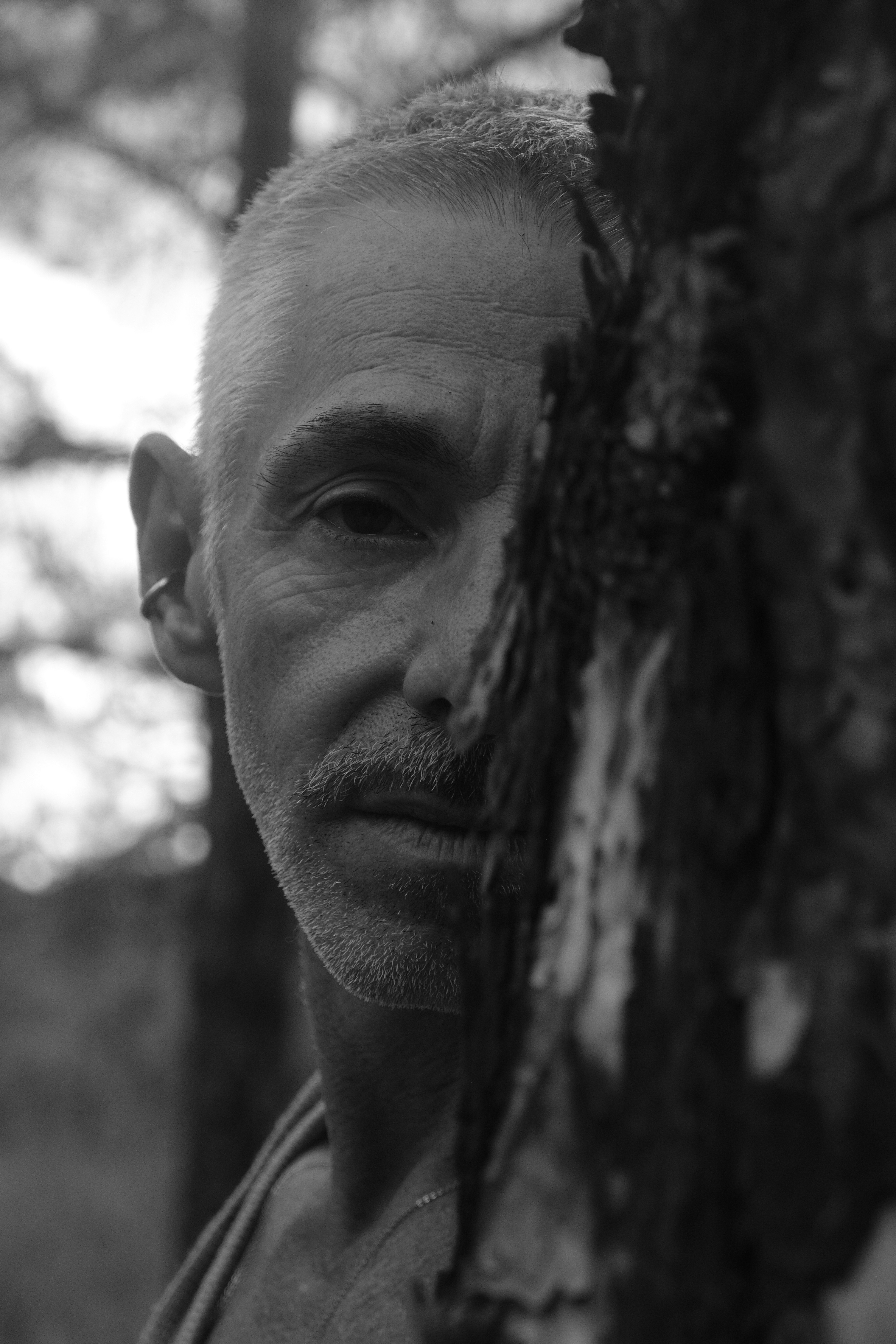A man with short grey hair and a mustache partially obscured by tree bark