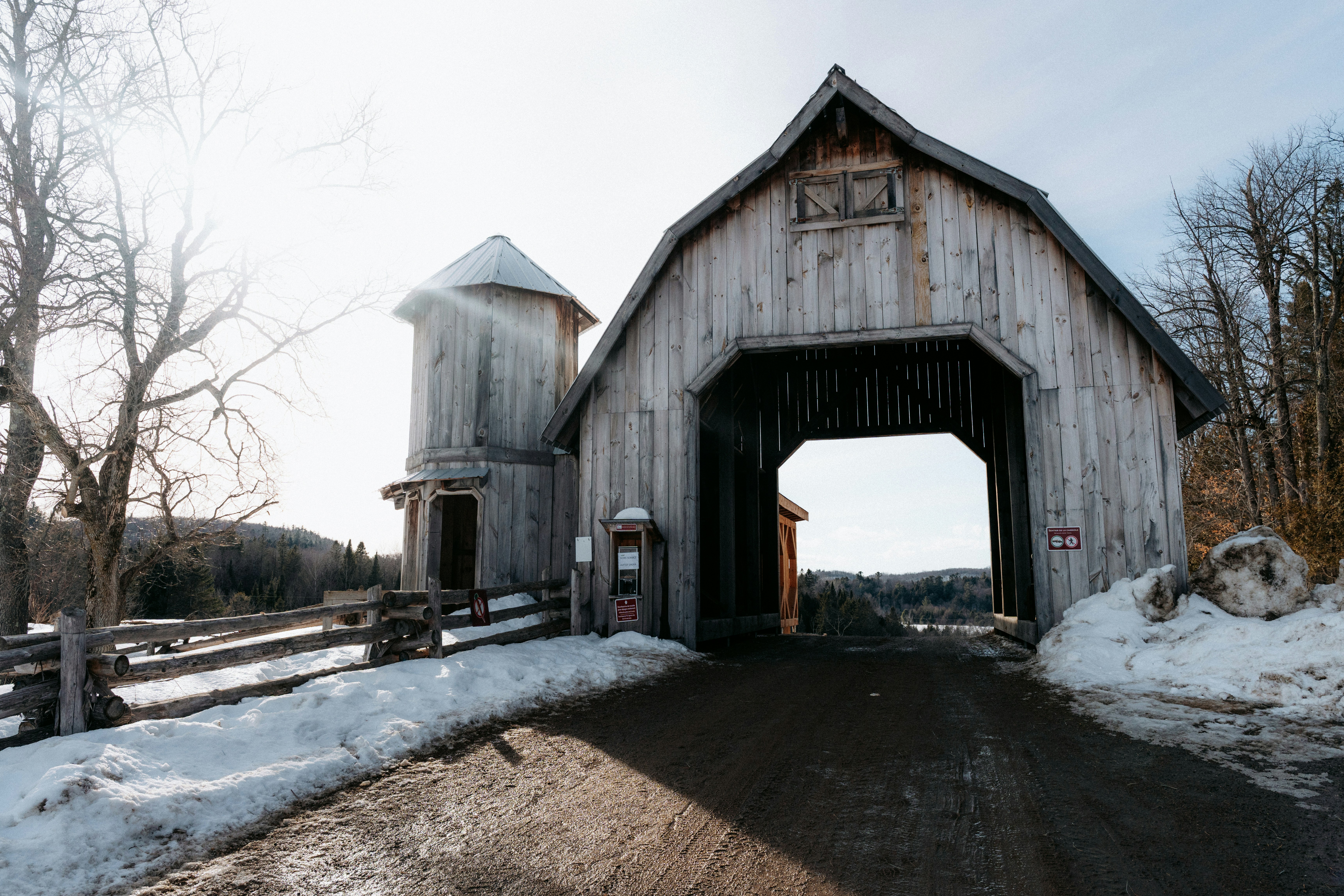 A wooden covered bridge with a silo in a snowy landscape under low sun