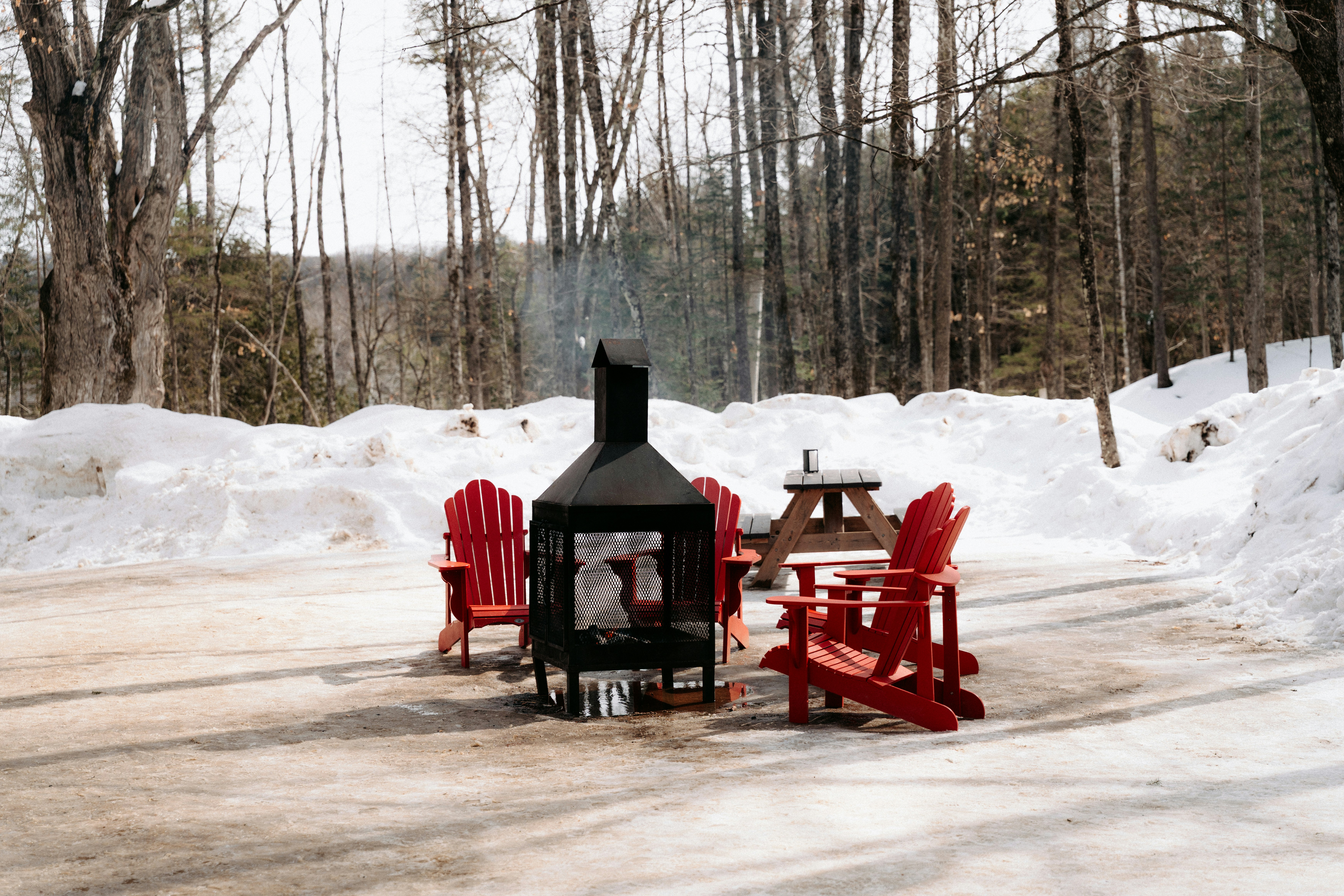 Red Adirondack chairs around a black metal fire pit in a snowy forest