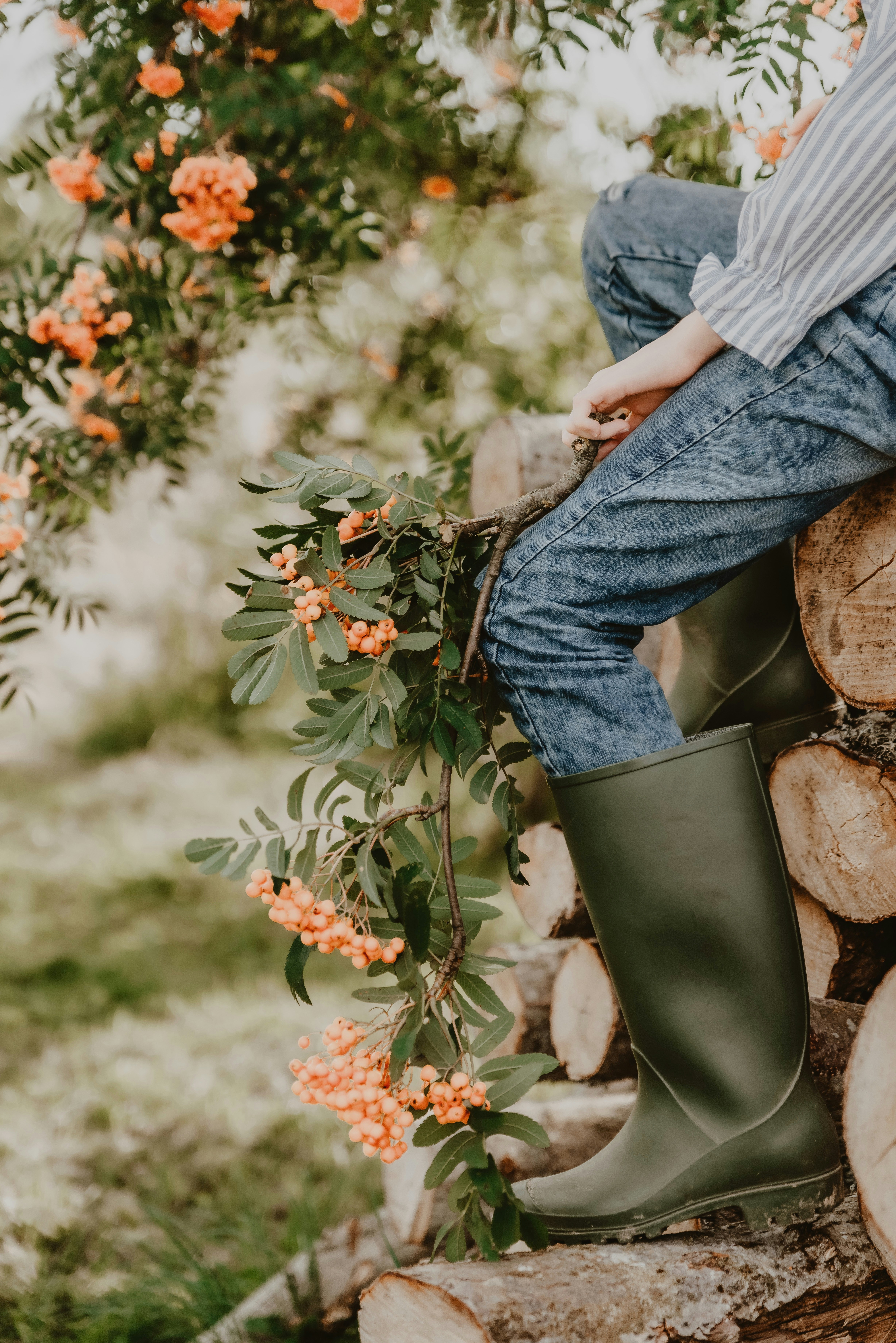 A person in blue jeans and green rubber boots sitting on stacked logs