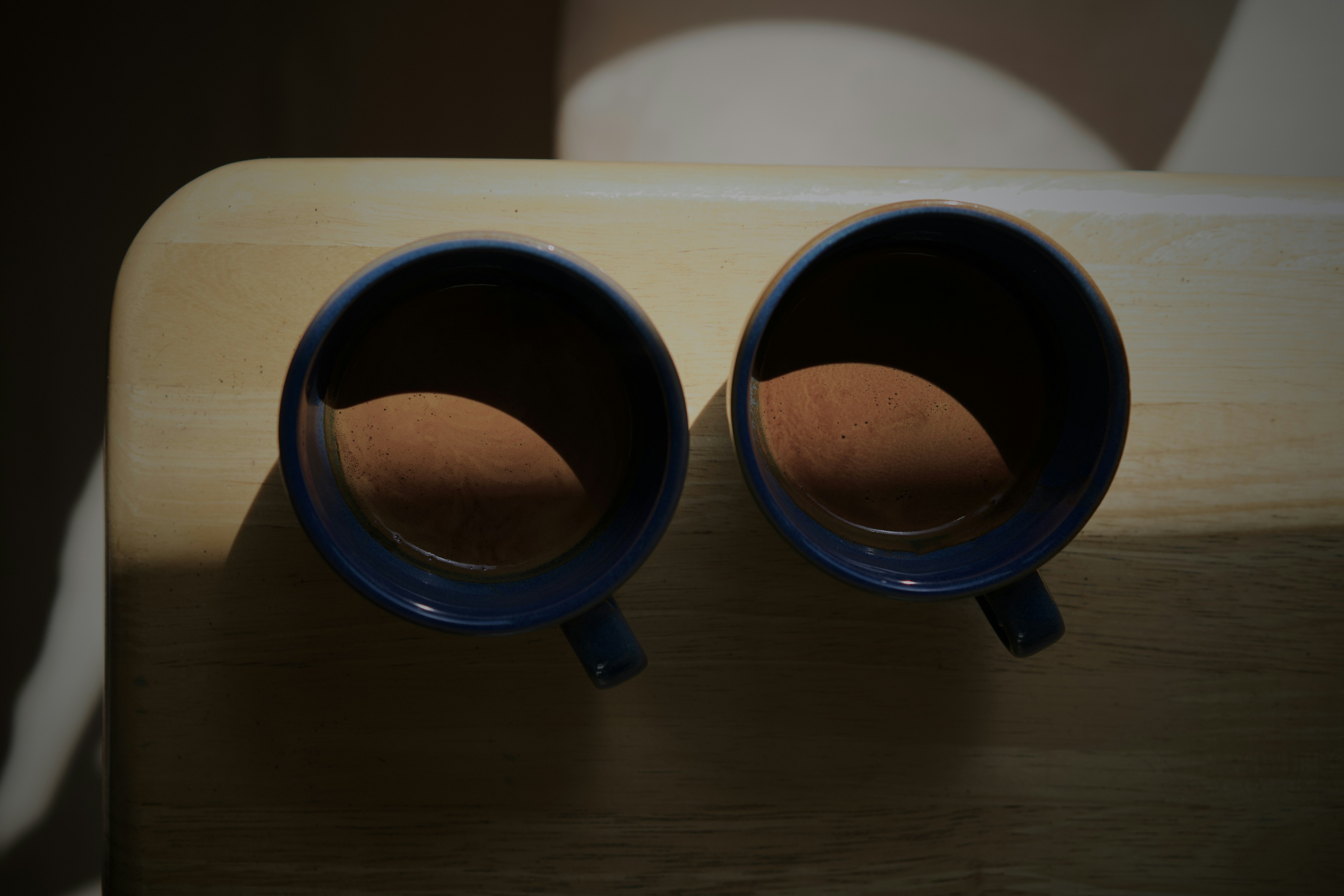 Two blue mugs filled with coffee on a light wooden table in sunlight