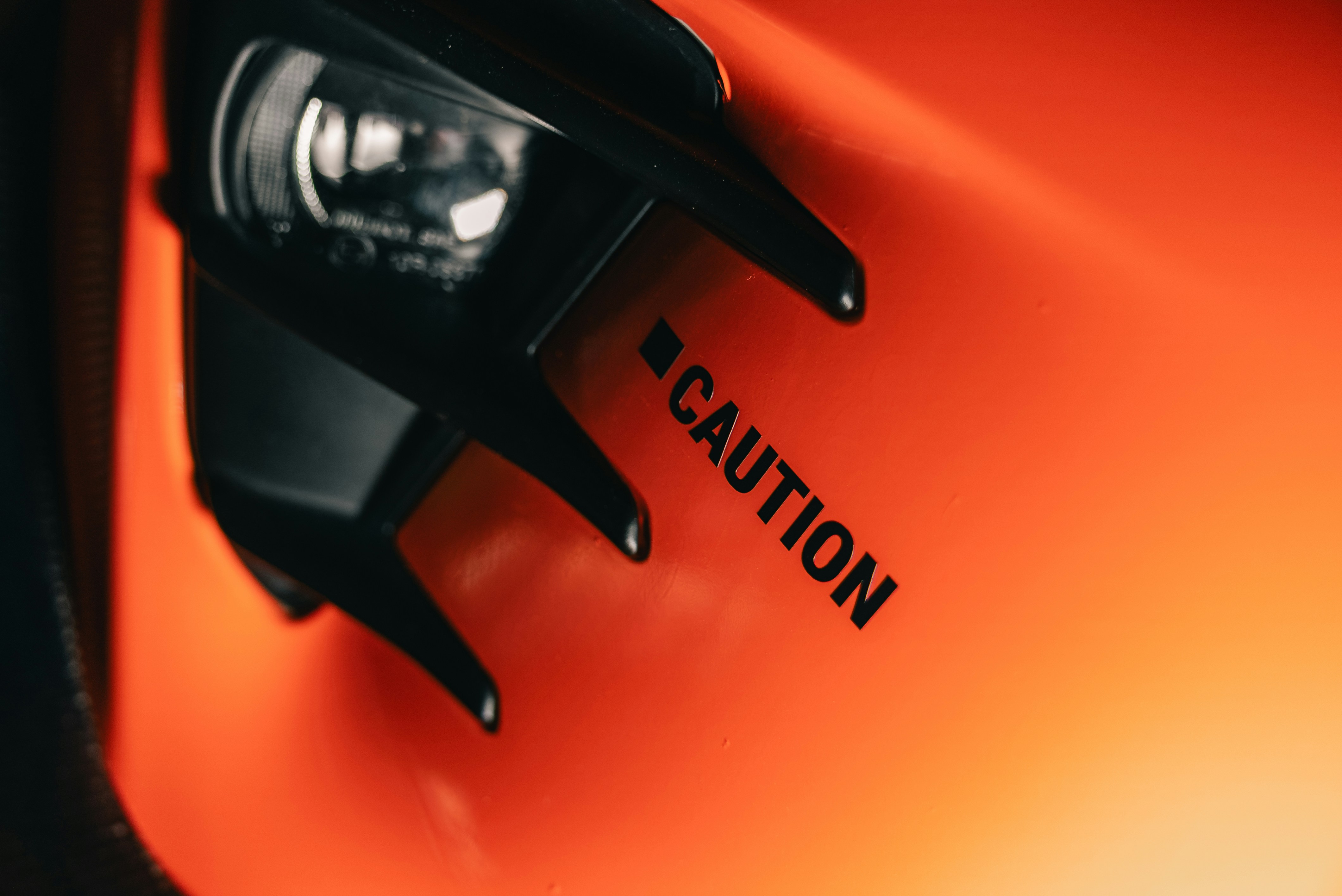 A black CAUTION sign on a bright orange surface near a dark headlight