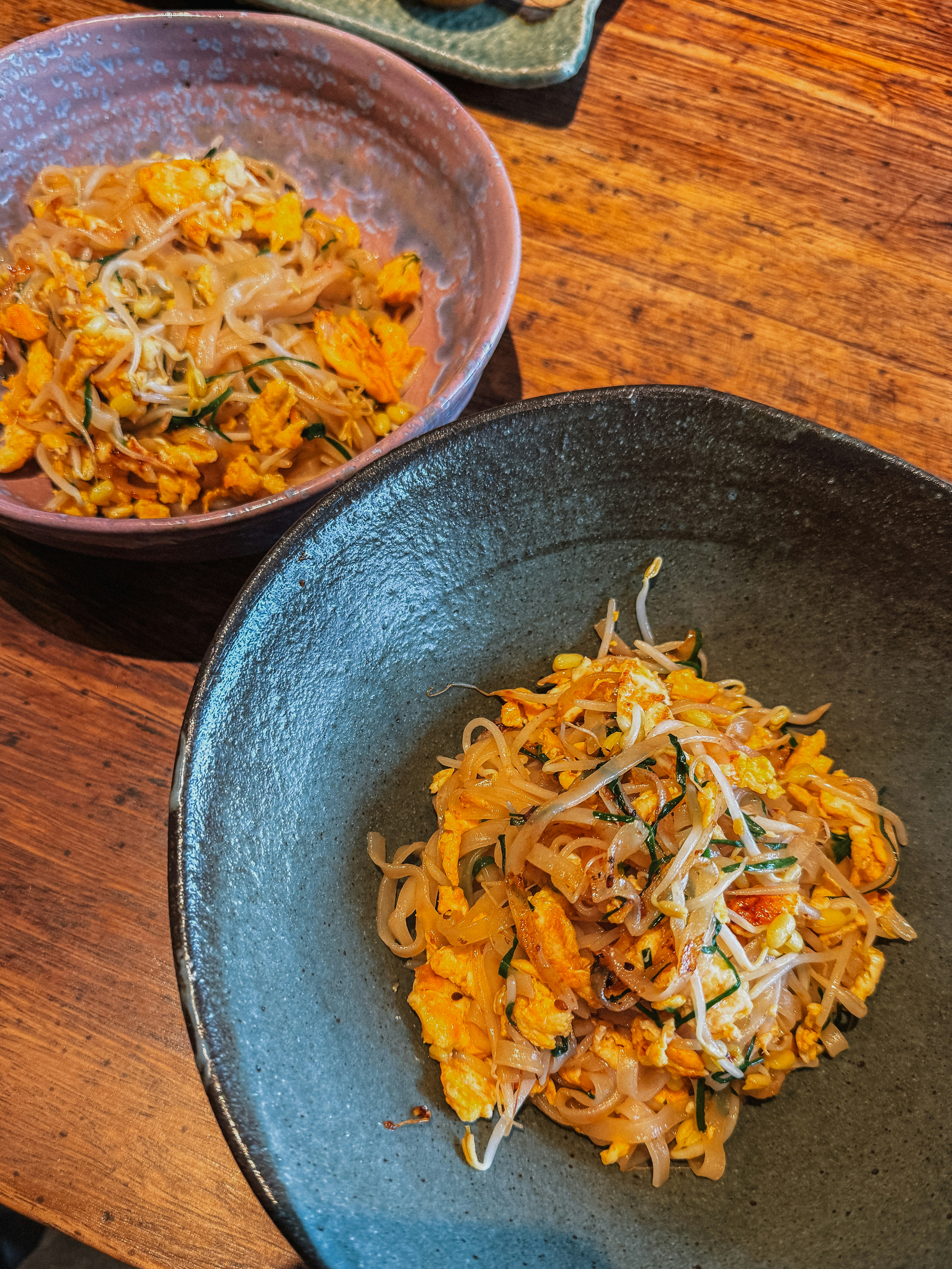 Two bowls of pad thai with scrambled egg, bean sprouts and chives on a wooden table
