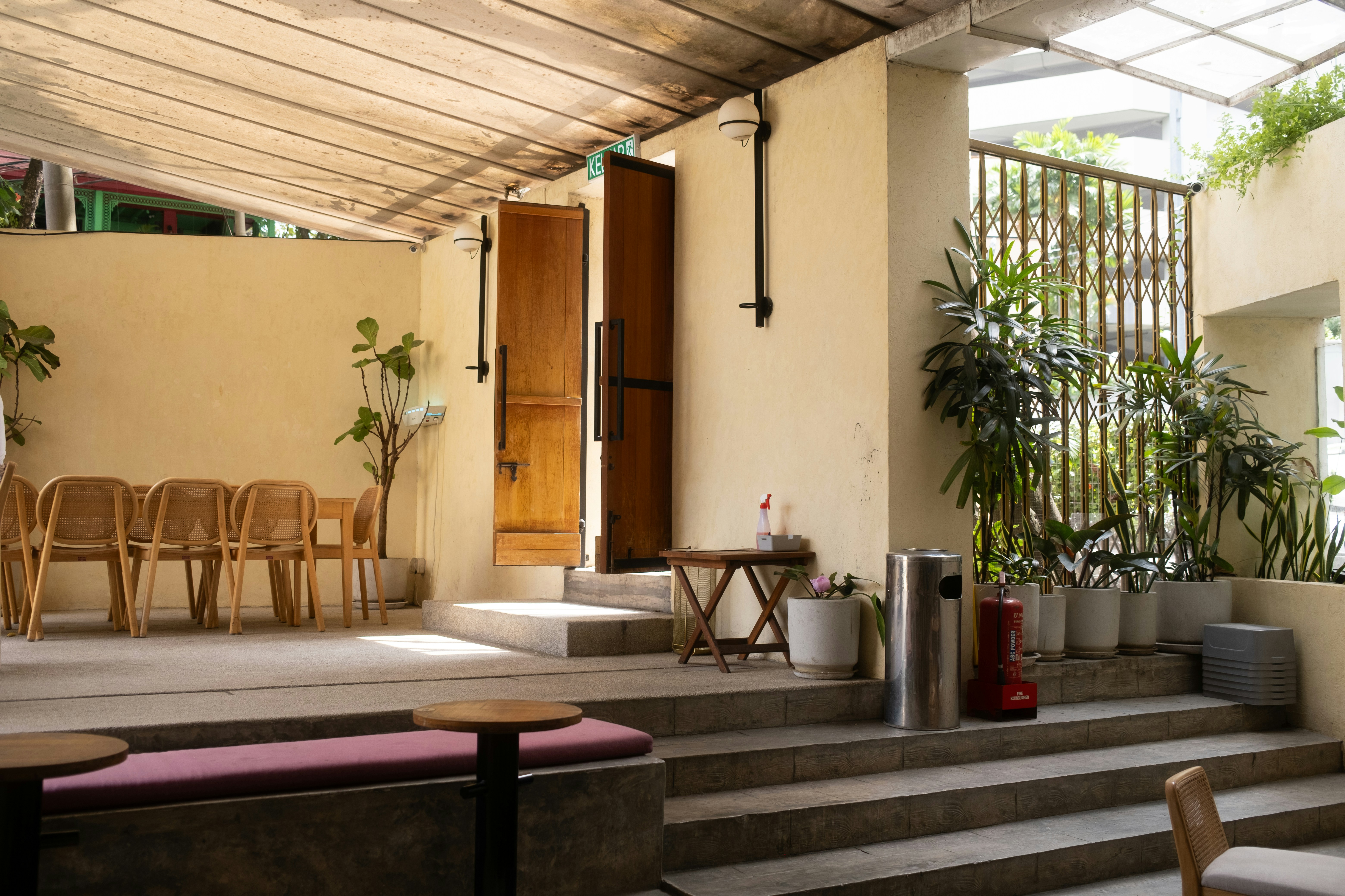 An open-air cafe terrace with rattan chairs, wooden doors, and potted plants