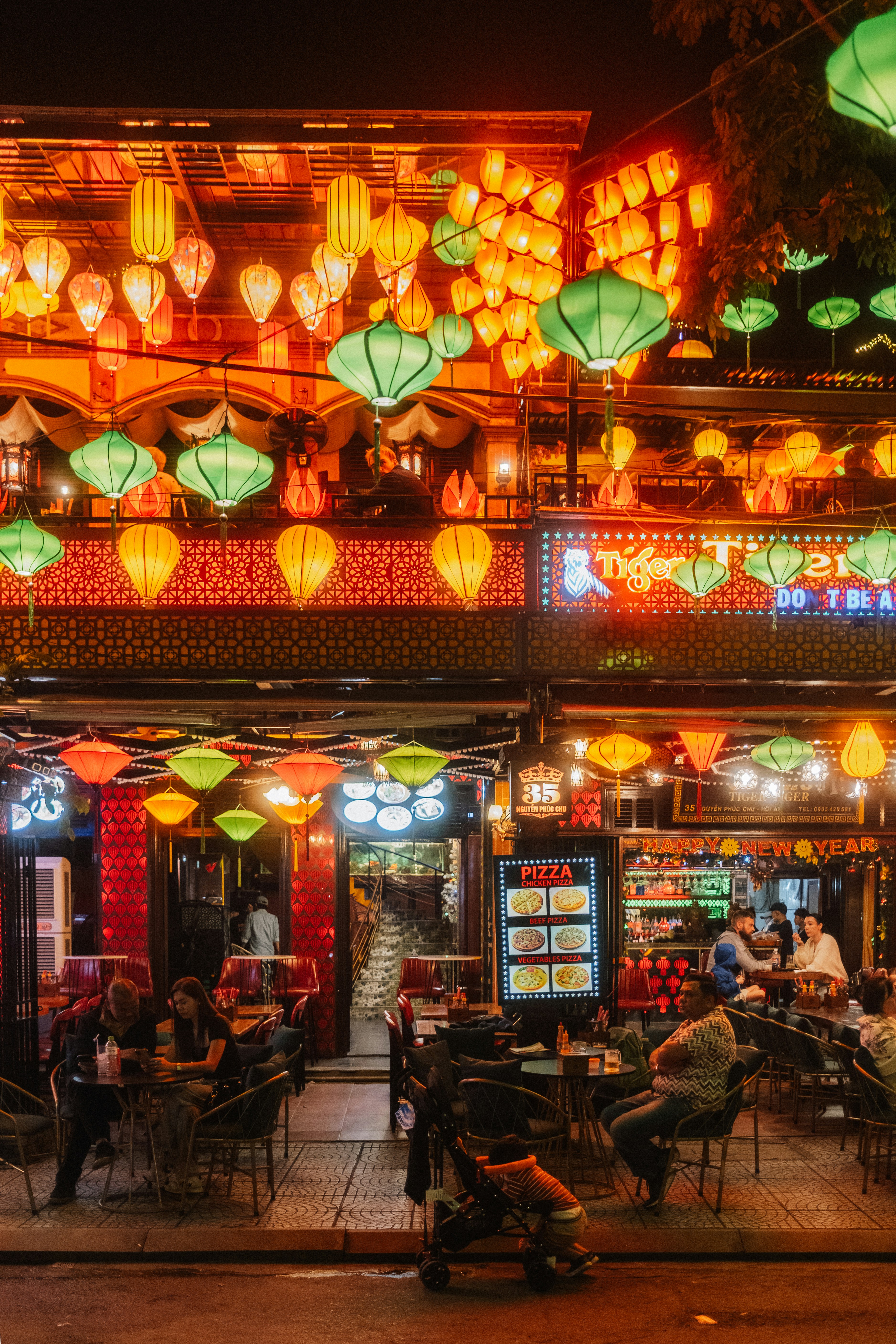 People dining at tables under colorful hanging lanterns on a street in Hoi An