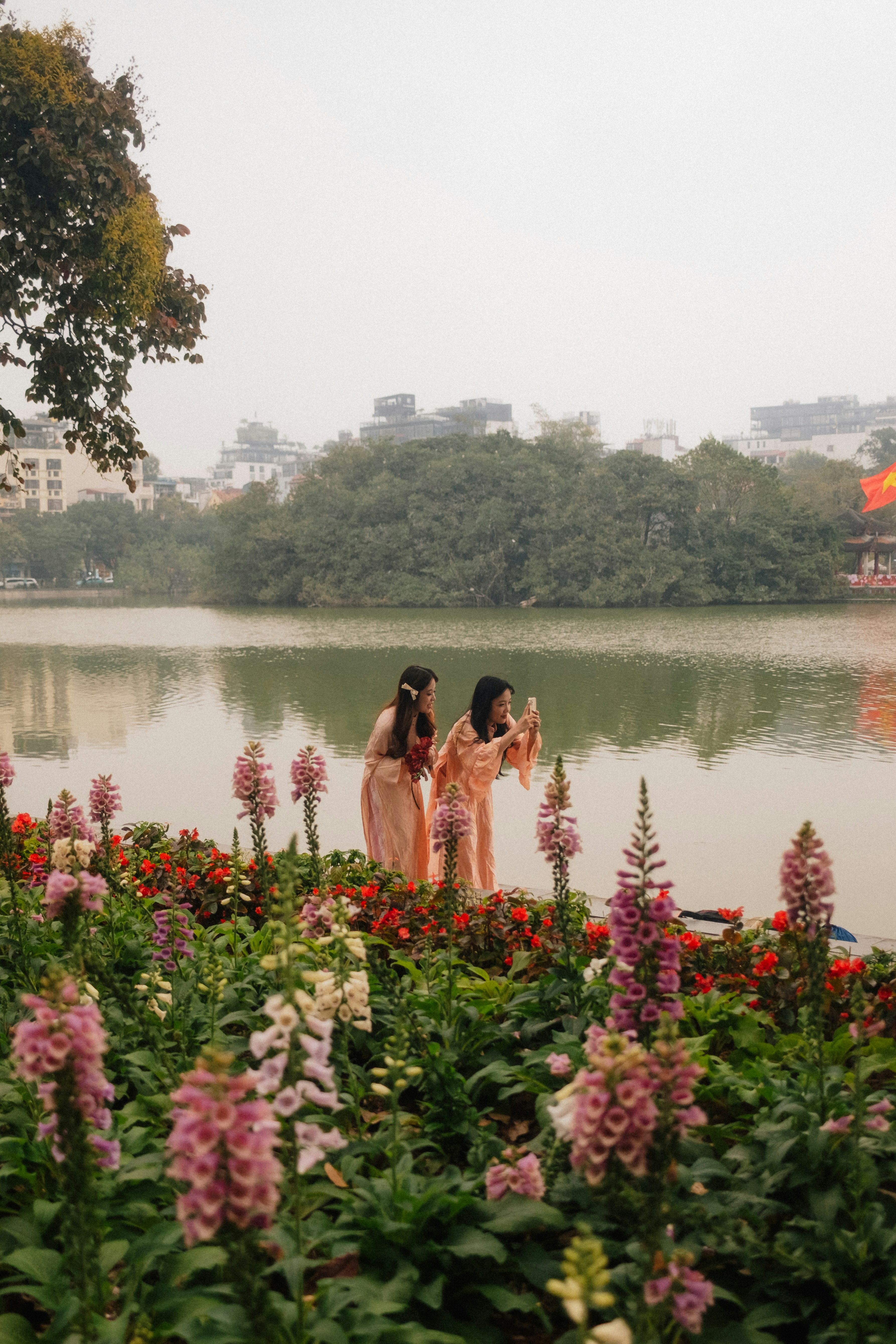 Photo of Two women in matching pink dresses taking a photo by a lake behind flower beds by Vincent Yap