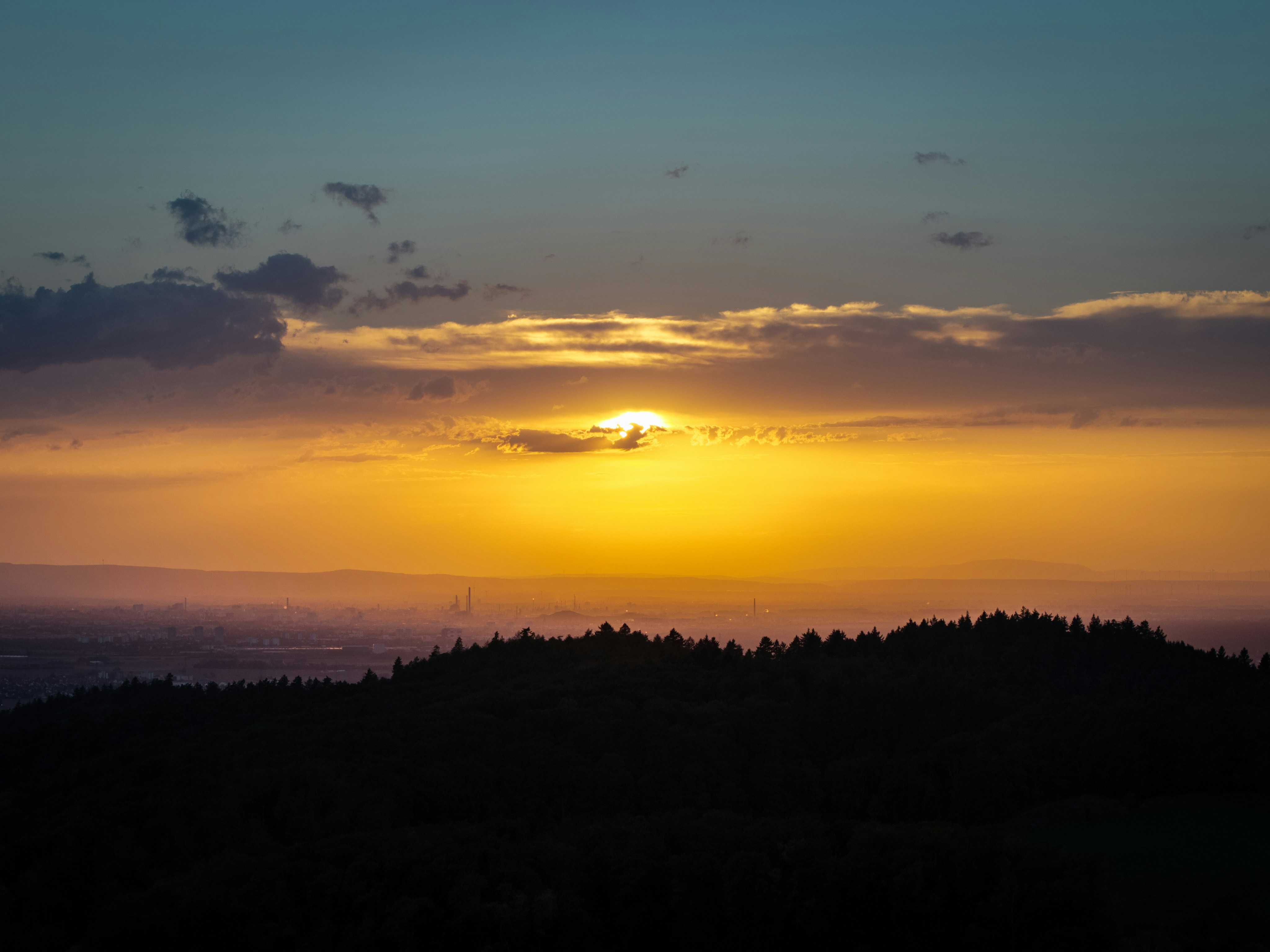 Low sun glowing behind clouds above a dark forested ridge and distant hazy city