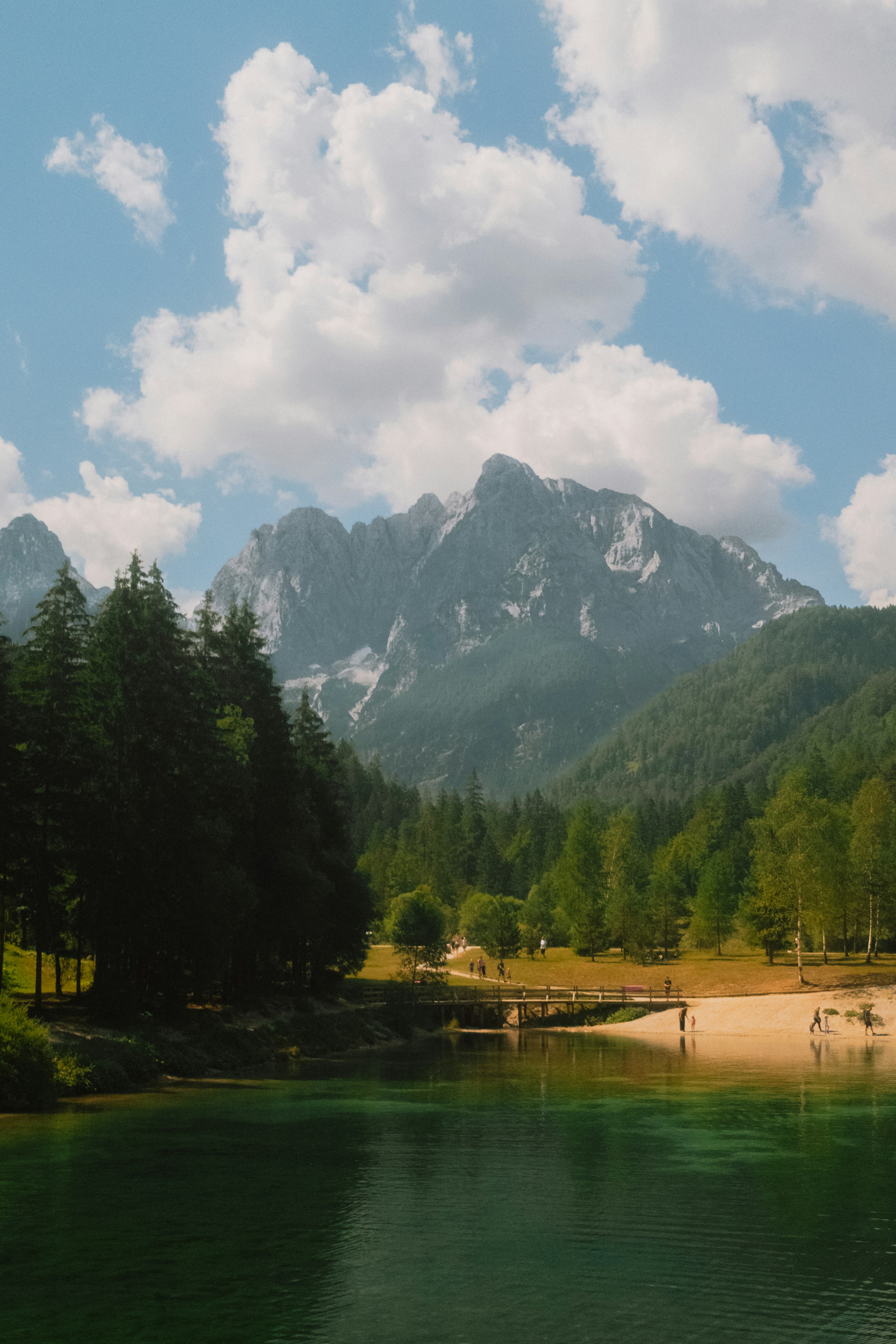 A wooden bridge over a green lake with mountains and forest in the background