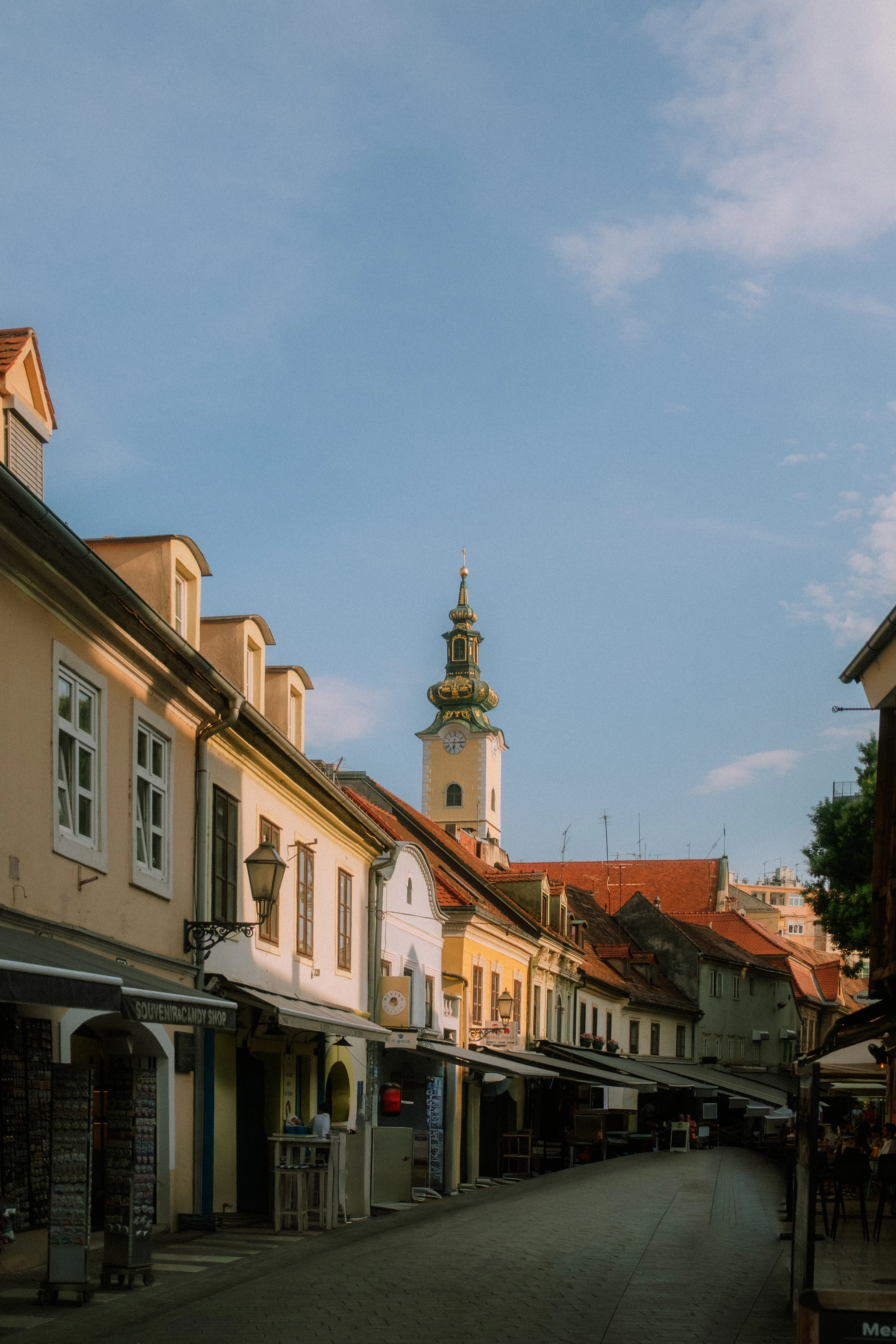 A cobblestone street glows in golden evening light, lined with shopfronts and awnings leading toward a church tower.