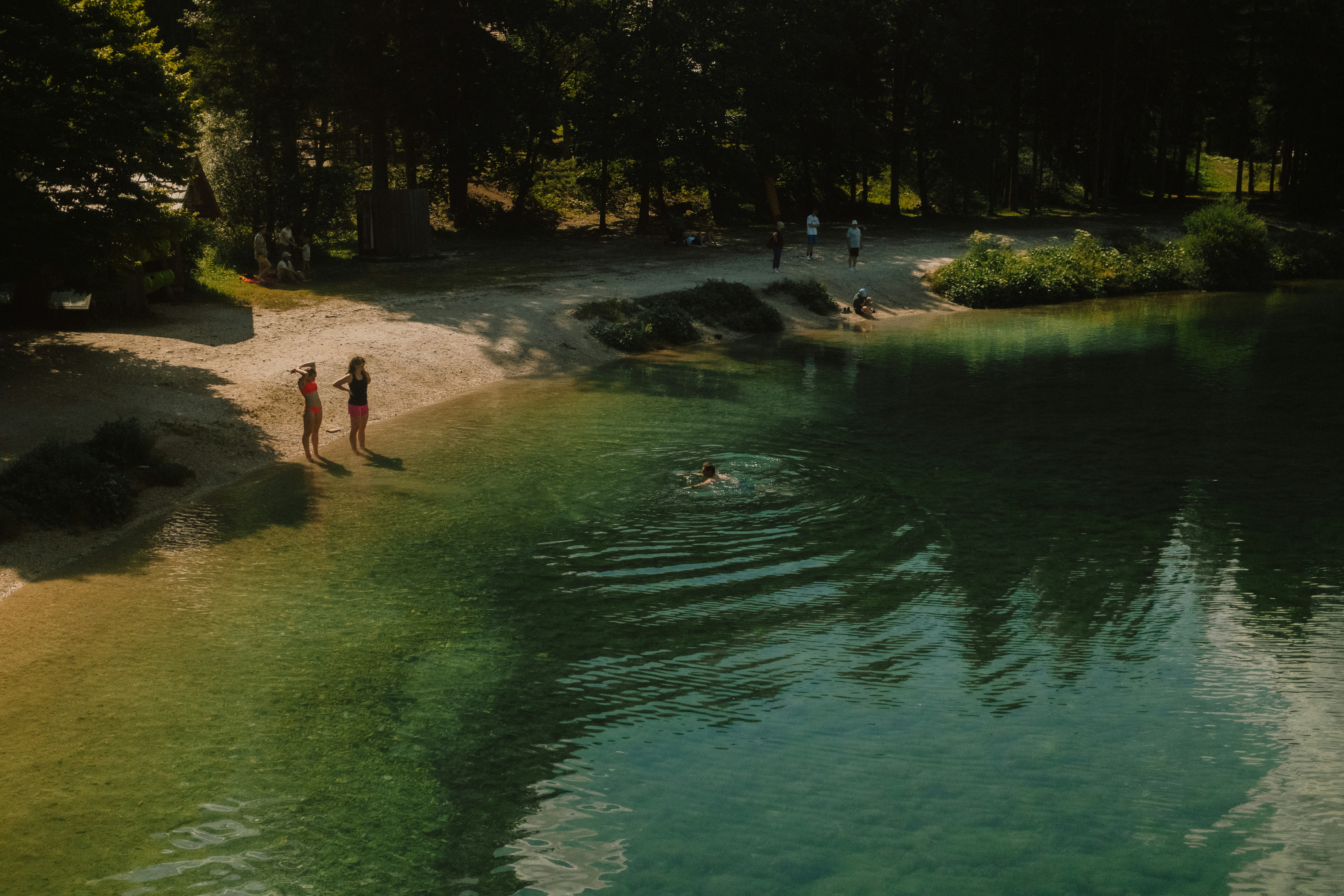 People on a sandy beach beside a clear green lake with trees