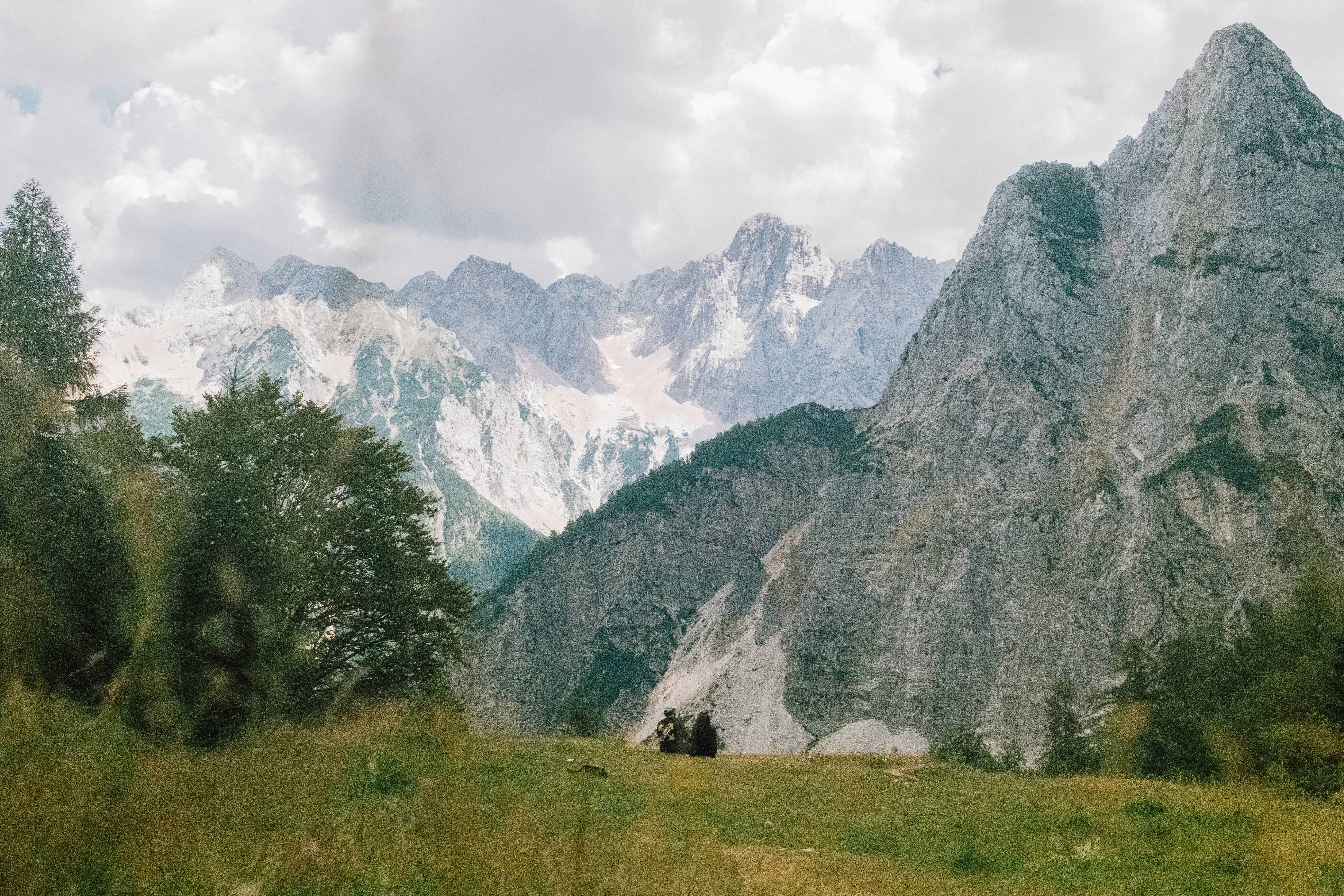 Two people sitting on a grassy hill before towering jagged mountain peaks