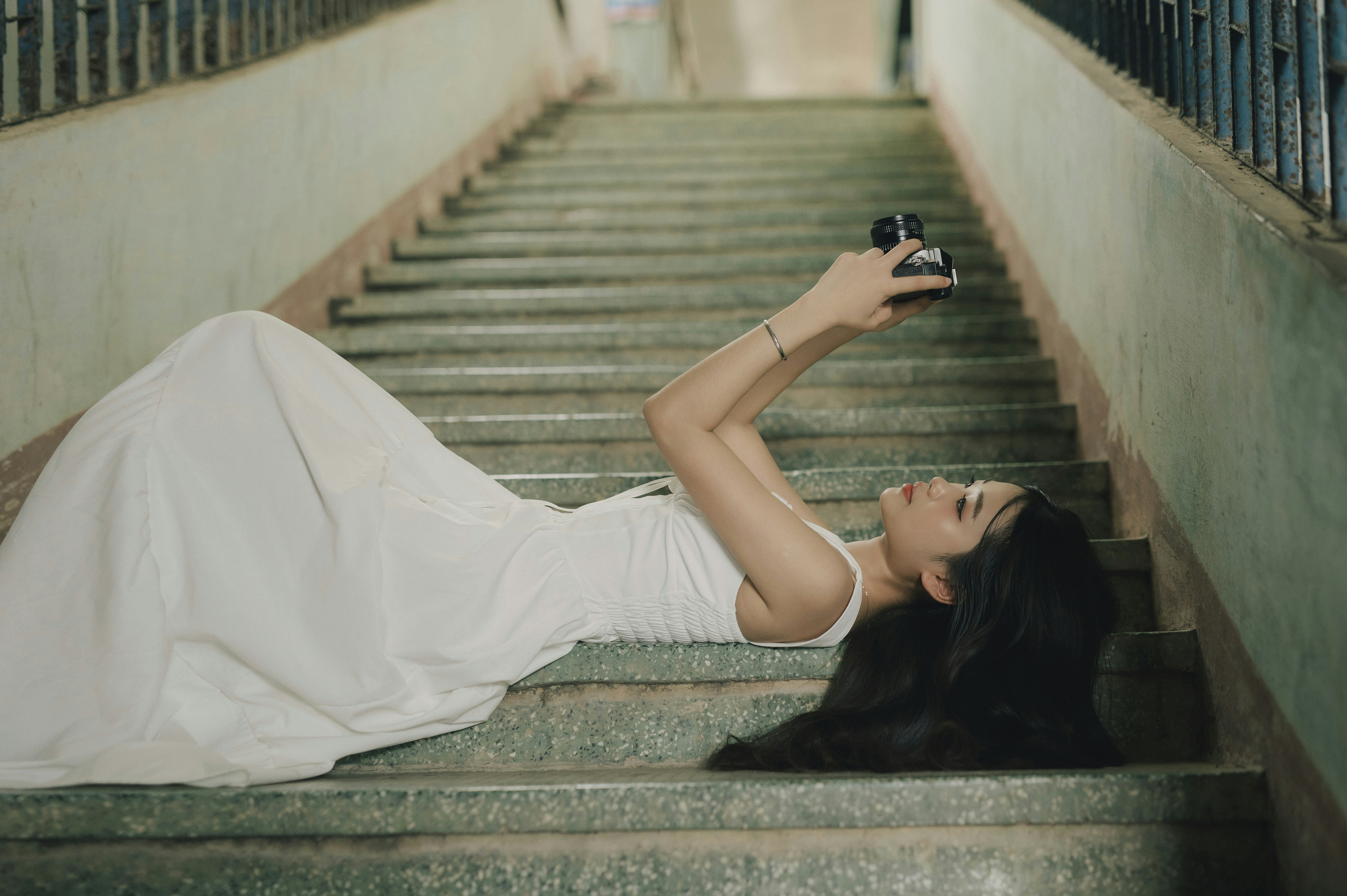 Photo of A woman in a white dress lying on concrete stairs holding a camera by Bin Thieu