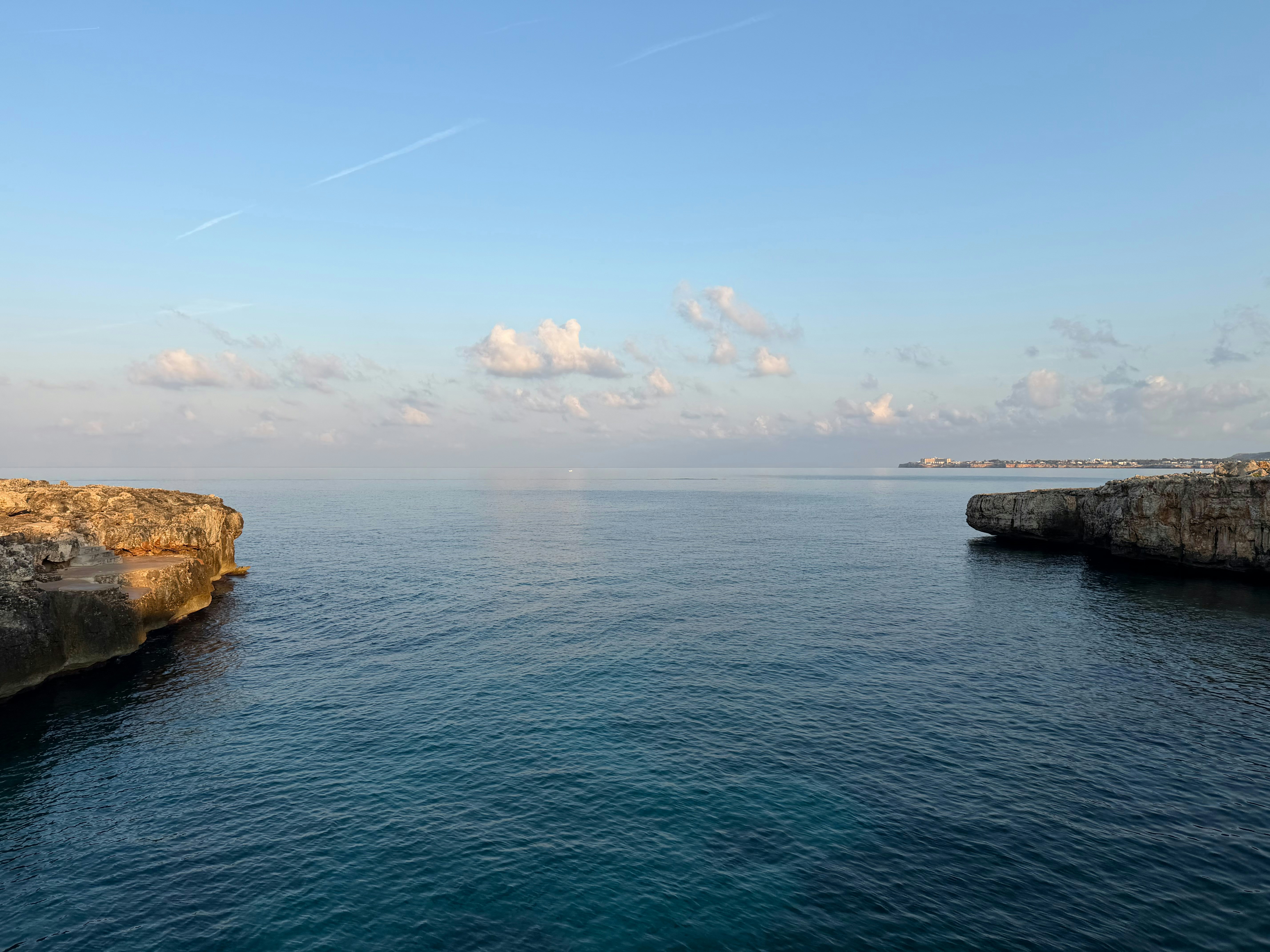 Calm blue sea between two rocky cliffs under a pale sky with clouds