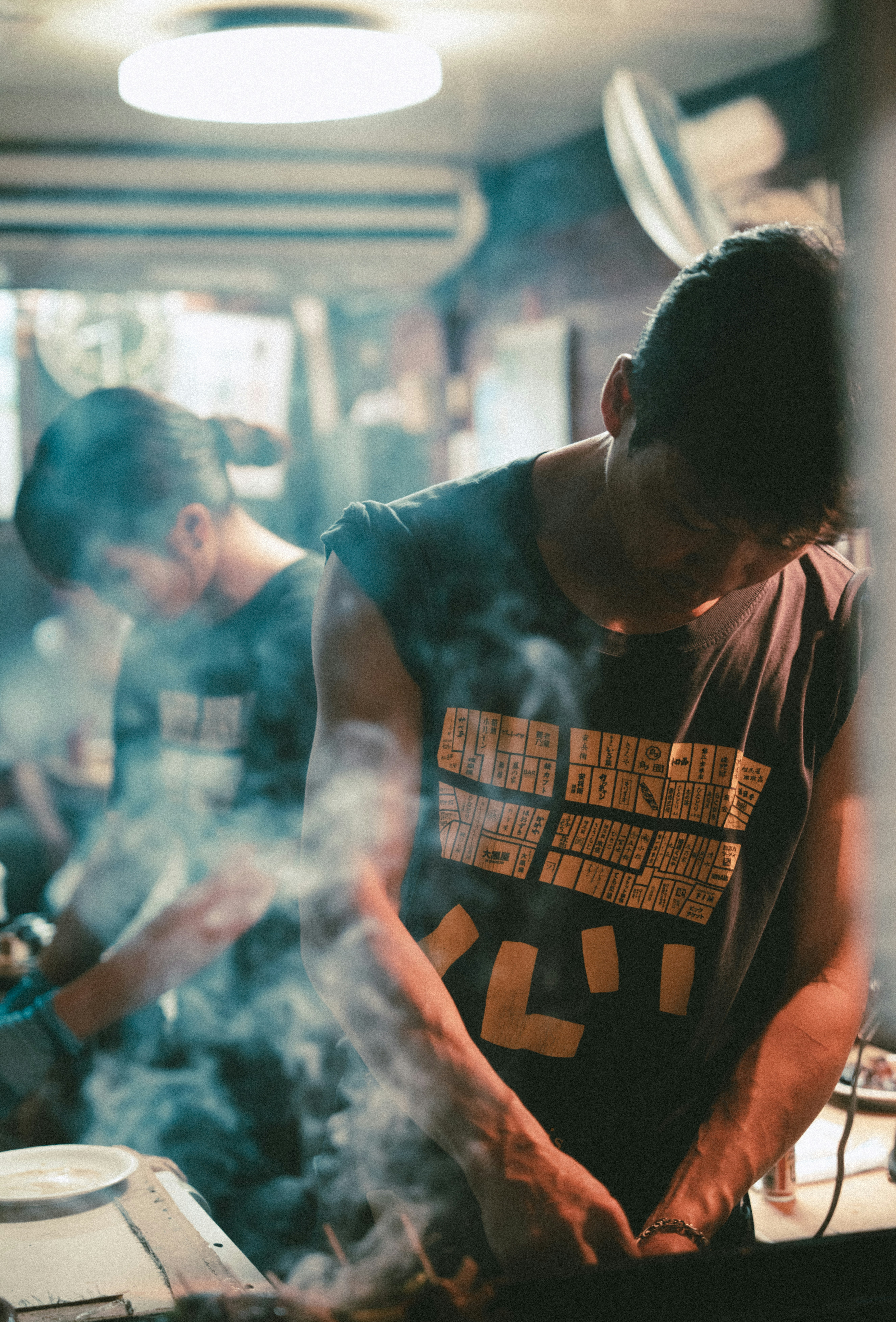 Two people in dark sleeveless shirts cooking food over a smoky grill