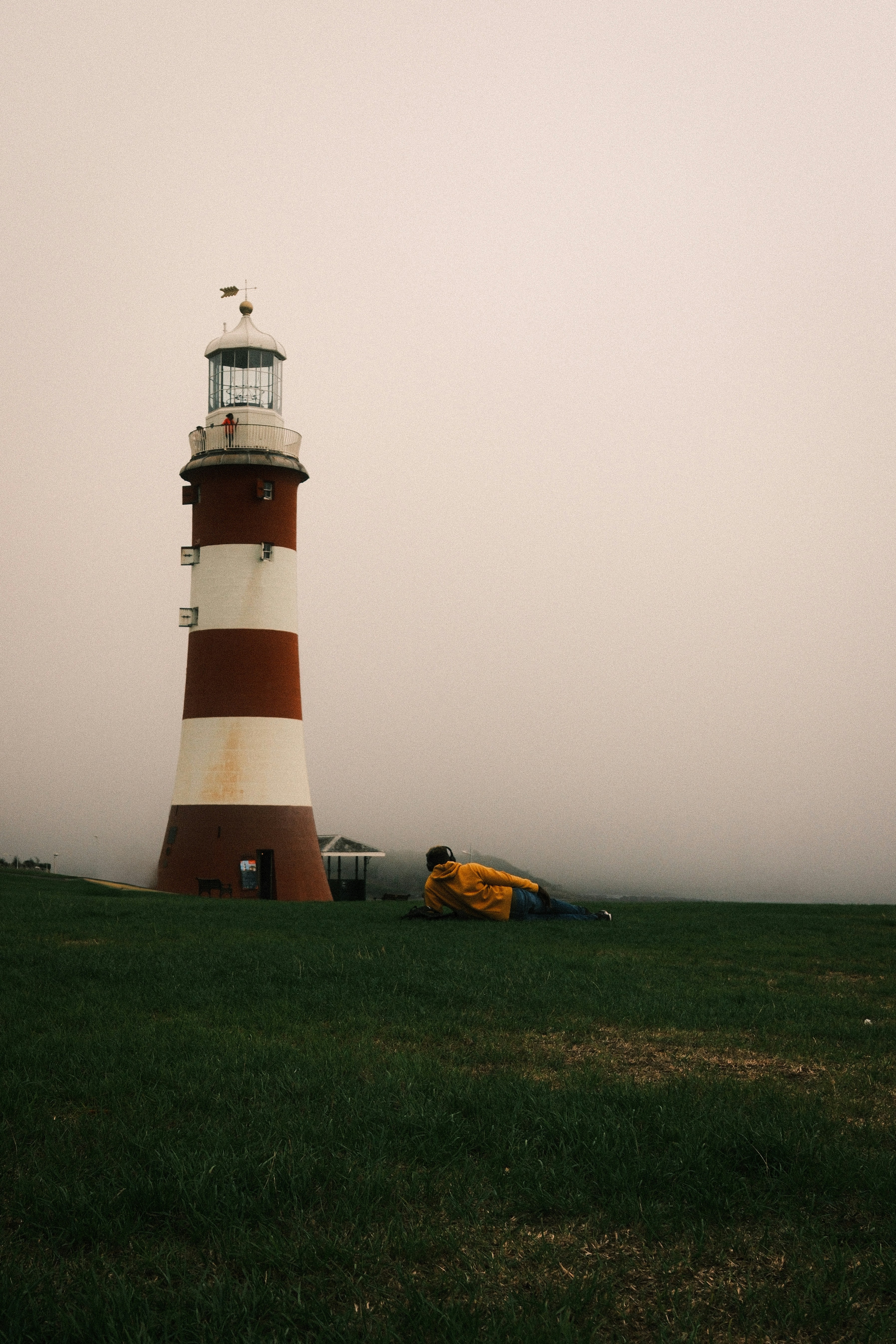 A person in a yellow hoodie lying on grass near a red and white lighthouse