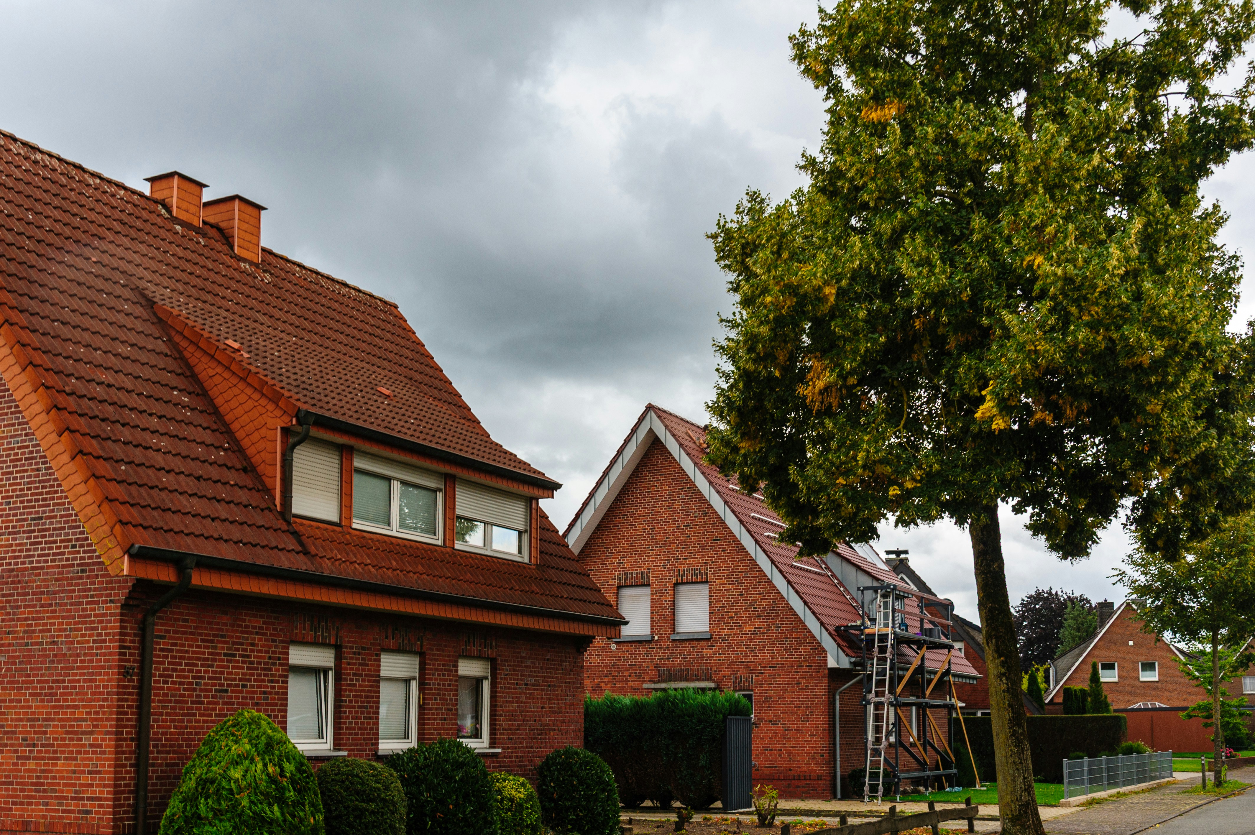 Red brick houses with tiled roofs under a cloudy sky near a large tree