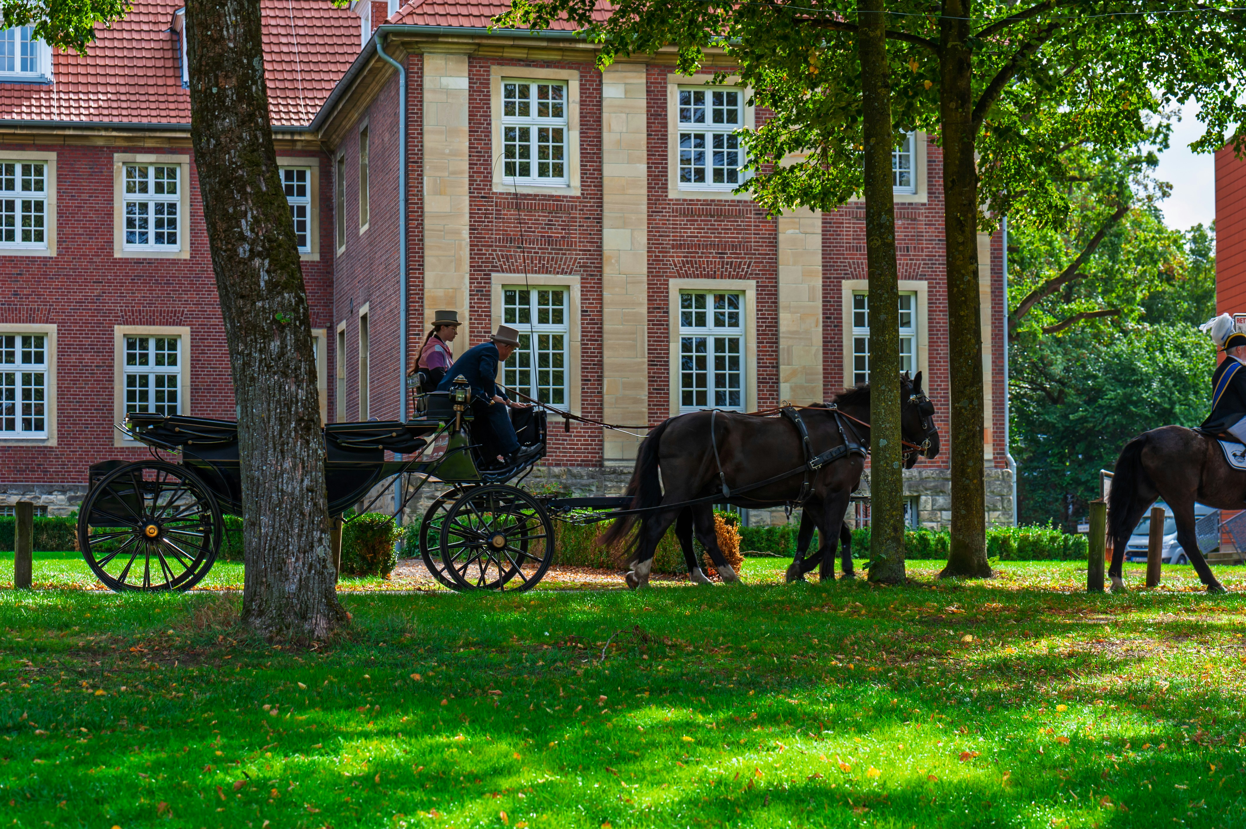 A horse-drawn carriage and a rider on horseback passing a brick building
