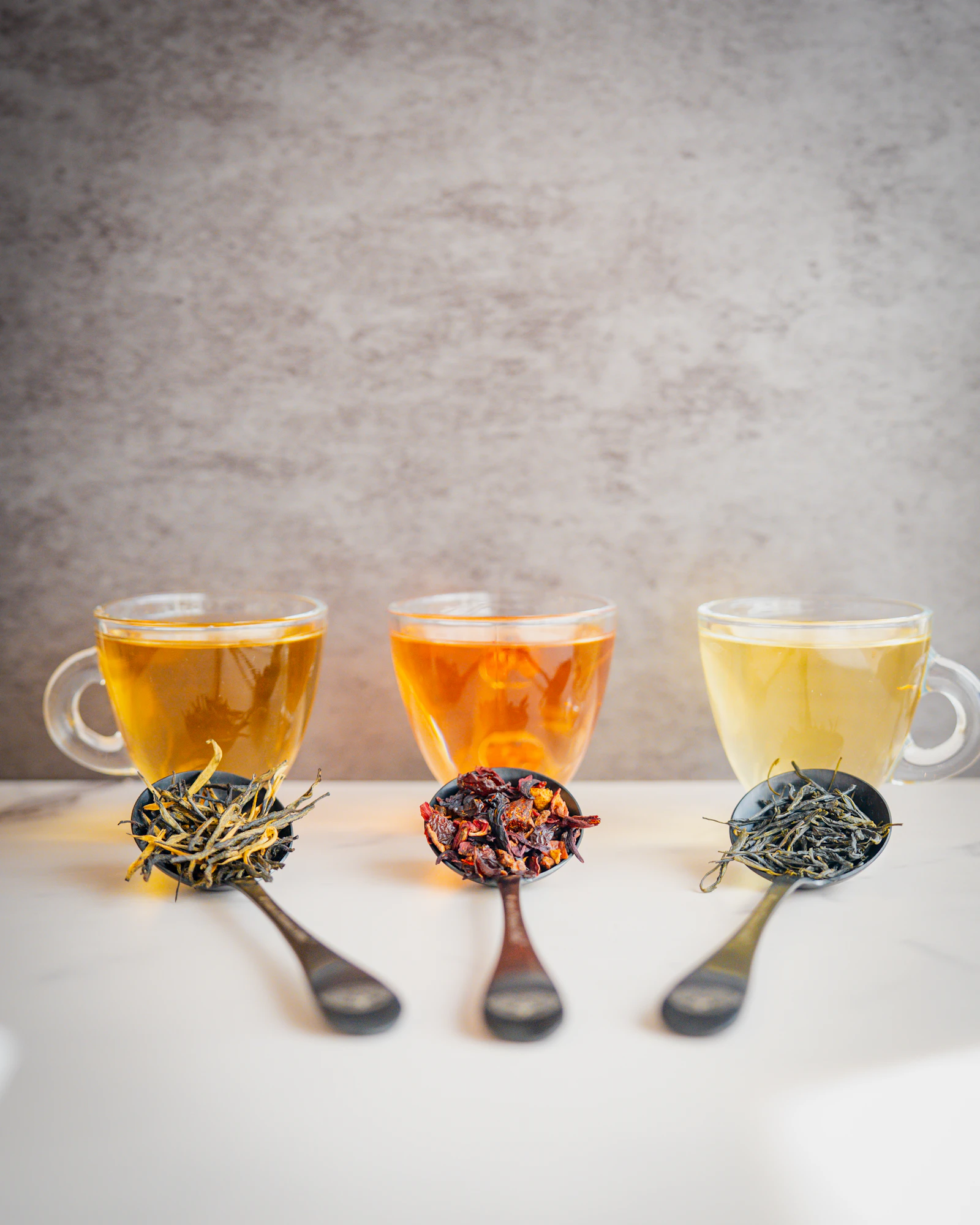 Three glass cups of brewed tea, each with a spoon of the loose leaves it was made from beside it