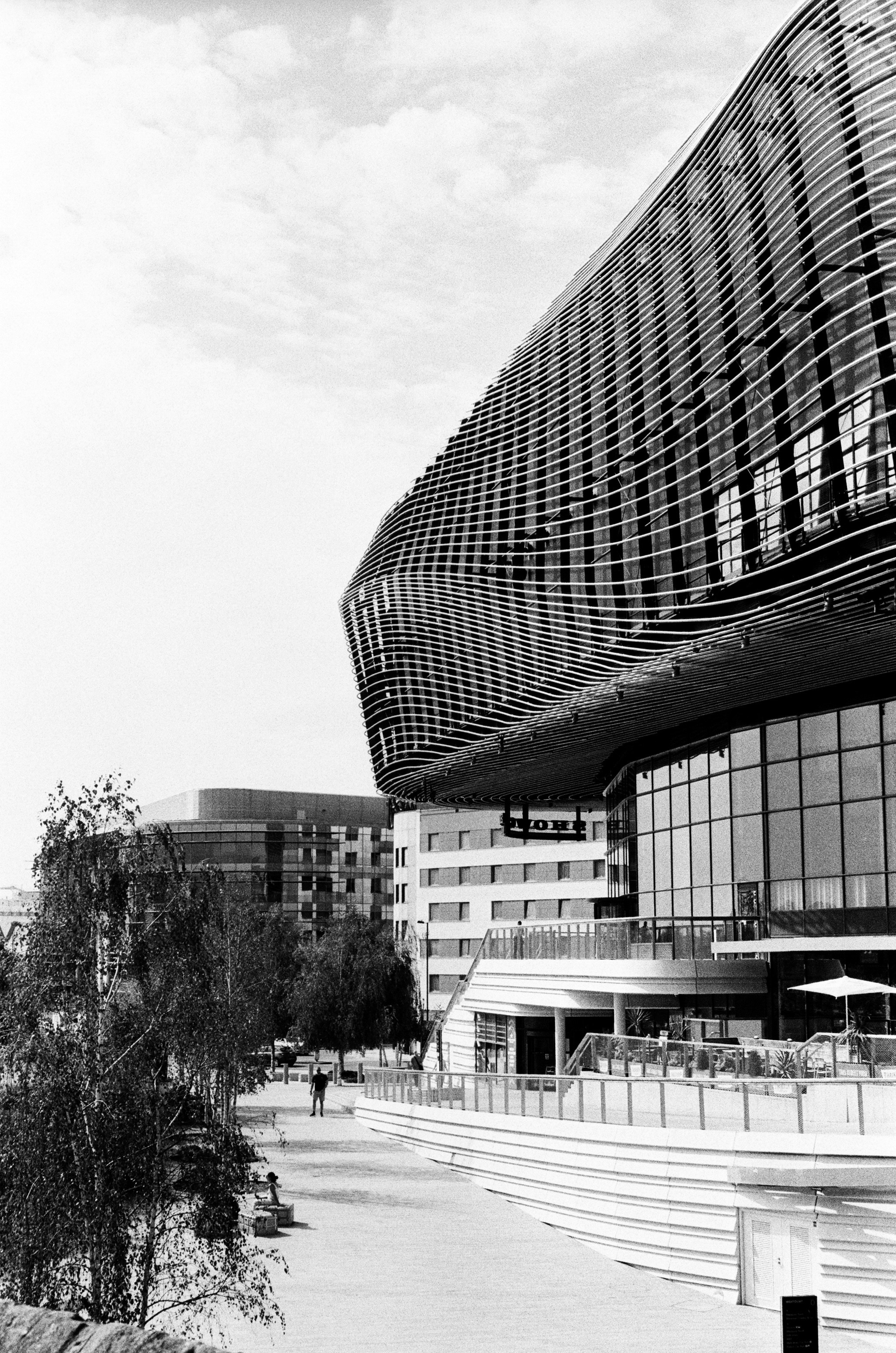 A modern building with curved metal louvers above a pedestrian walkway and trees