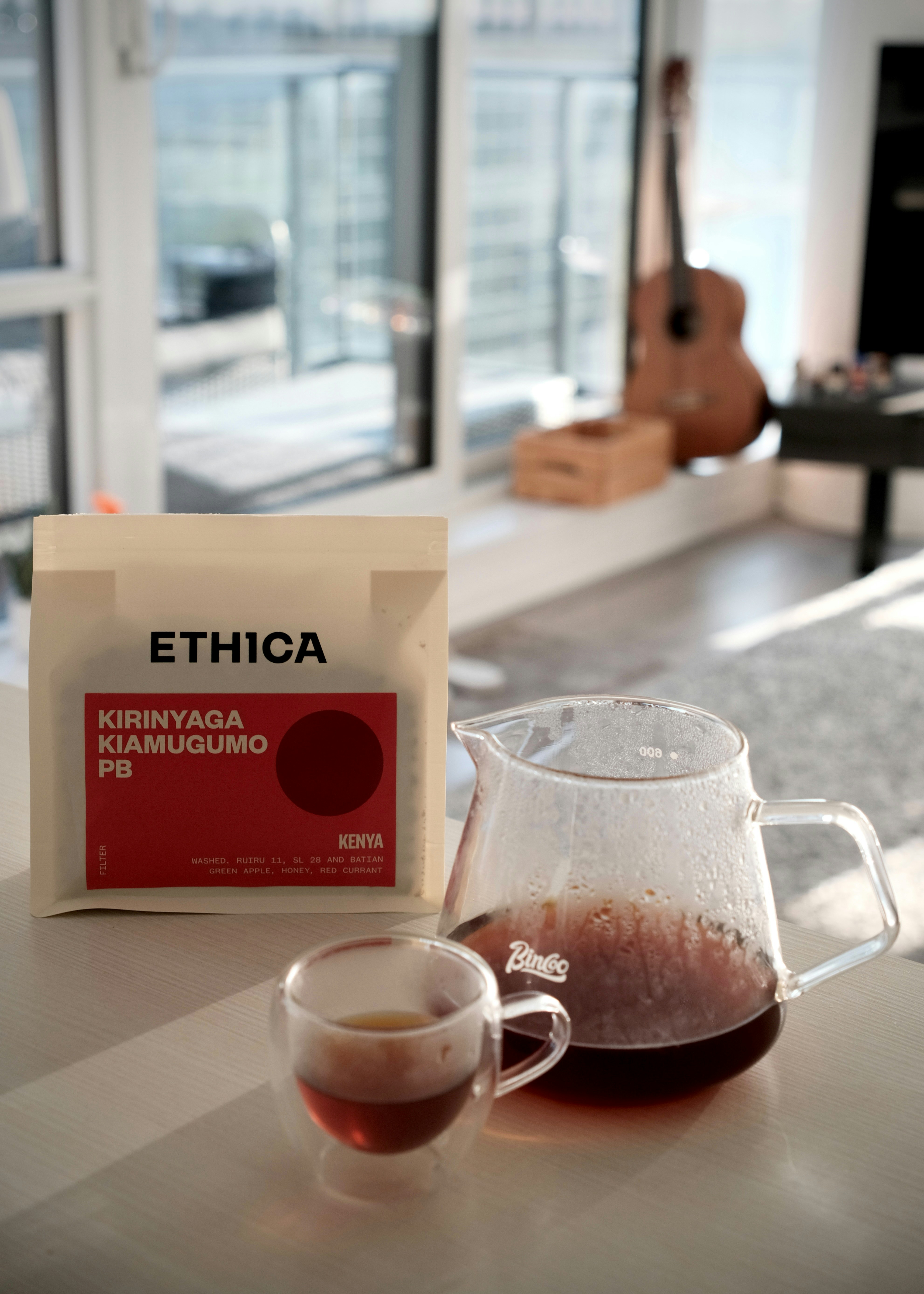 Sekantong kopi Ethica di samping sebuah karaf kaca dan cangkir di atas meja
