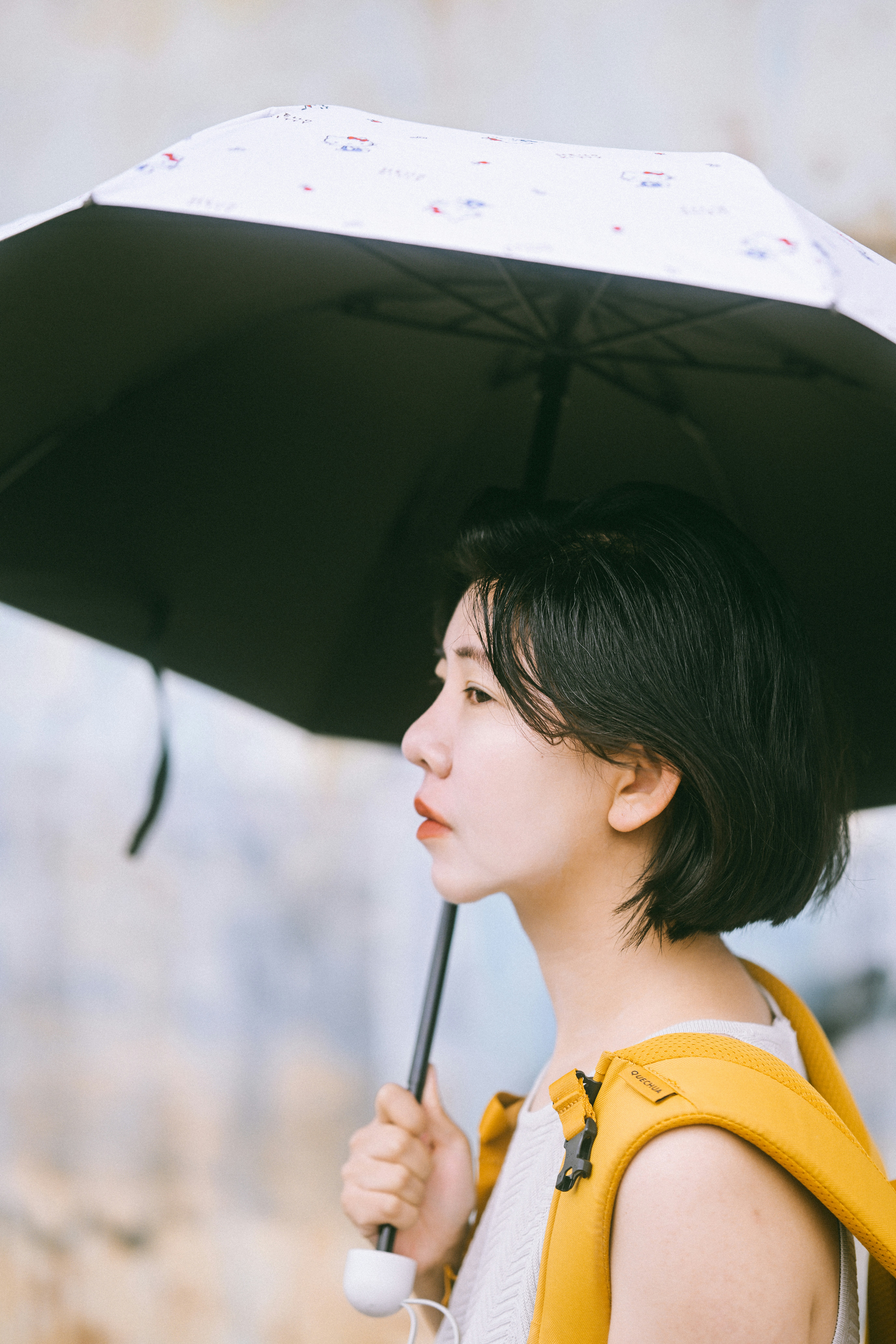 Une femme aux cheveux courts et foncés, tenant un parapluie noir et portant un sac à dos jaune