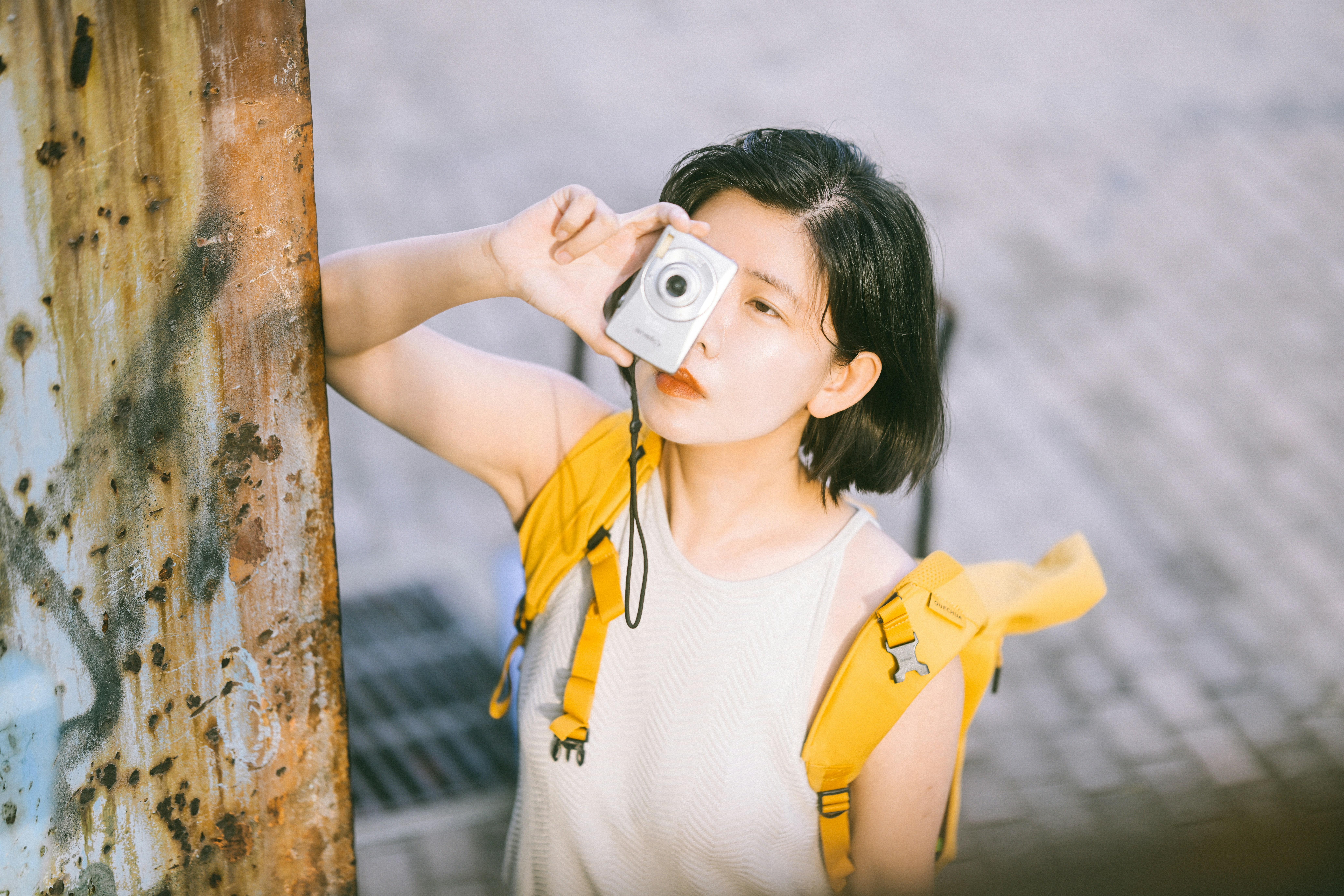 Photo of A woman with a yellow backpack holding a small silver camera to her eye by Sou Jest