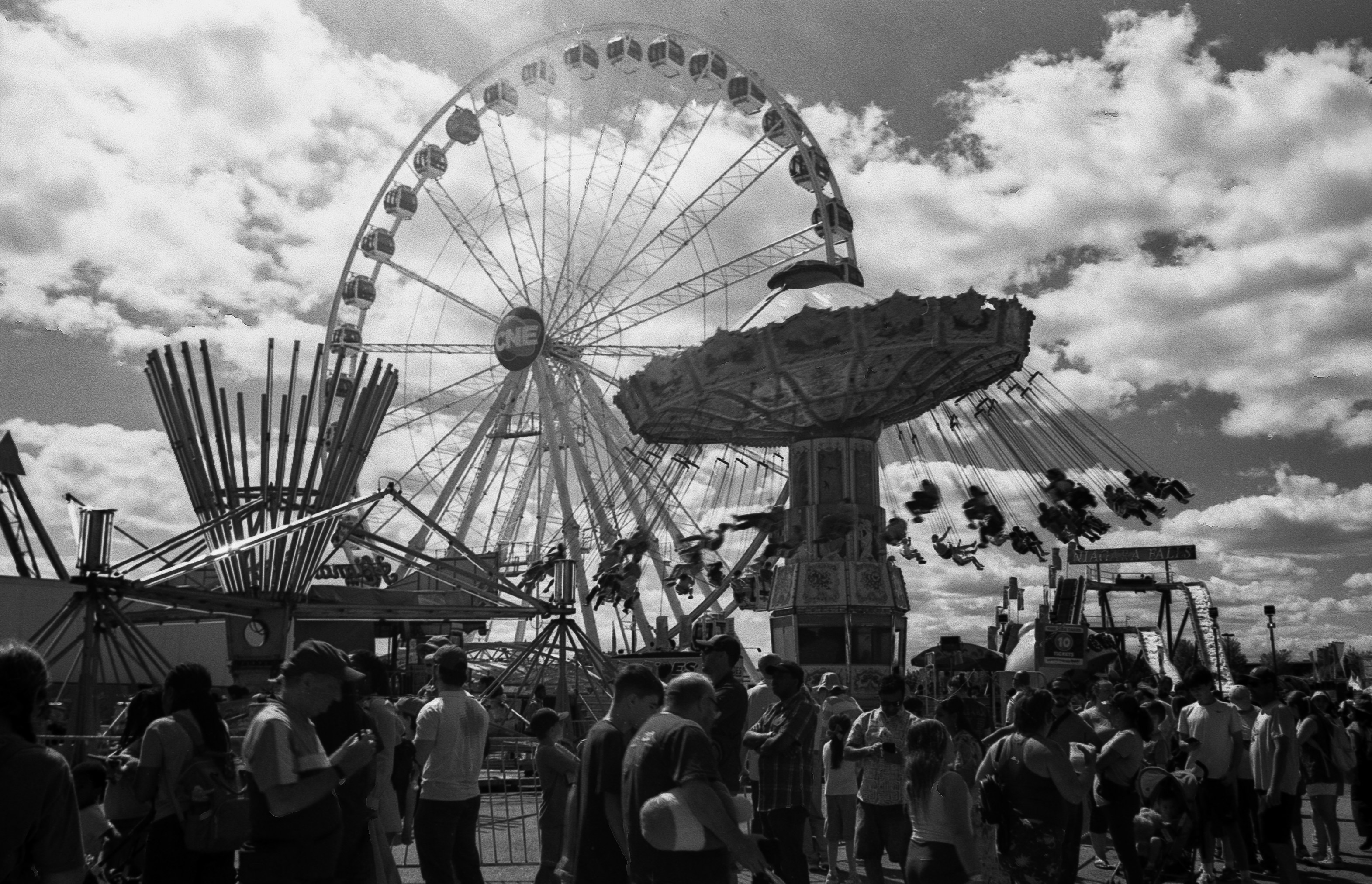 A crowd of people at a fairground with a ferris wheel and swing ride