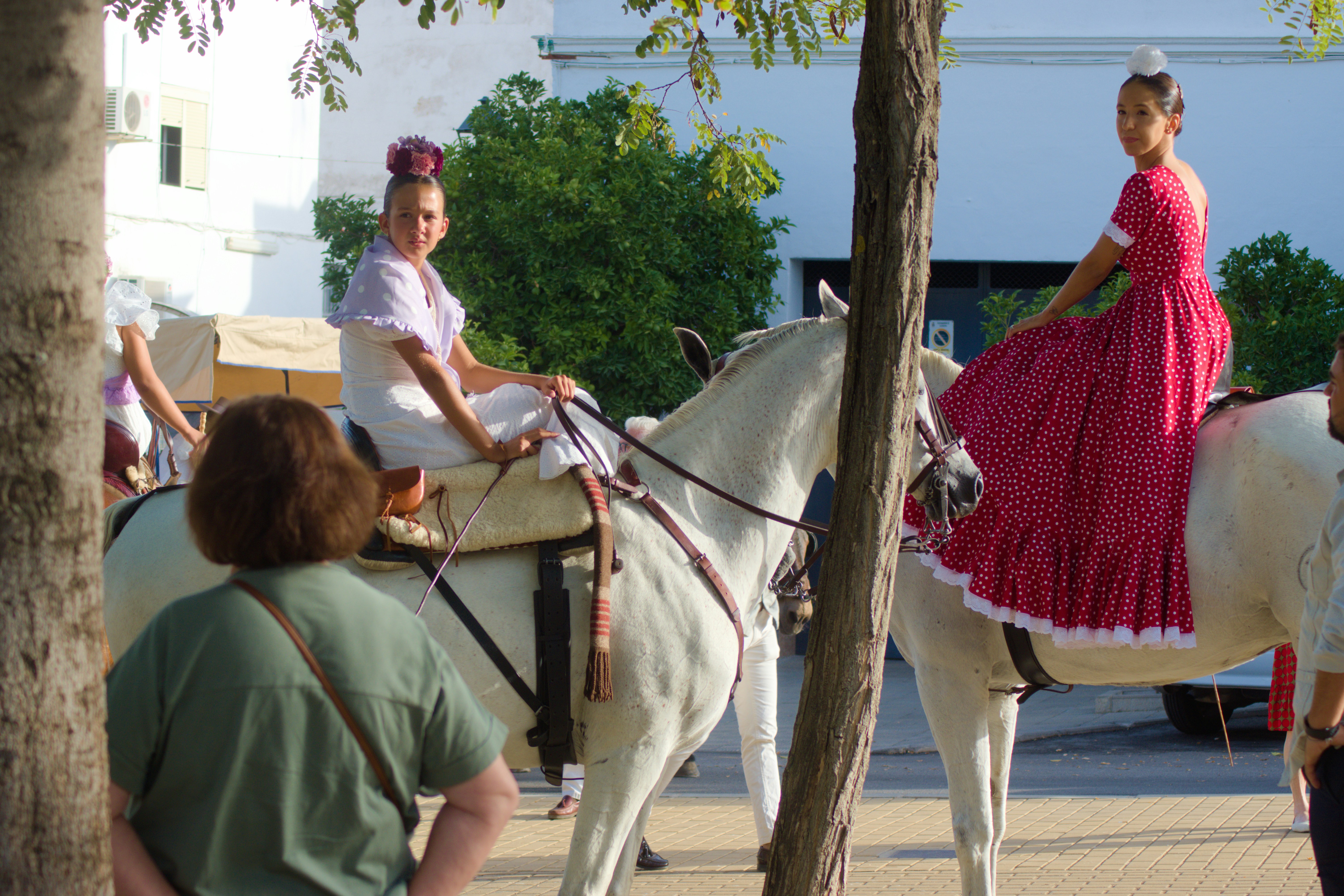 Two women in traditional flamenco dresses riding white horses on a paved street