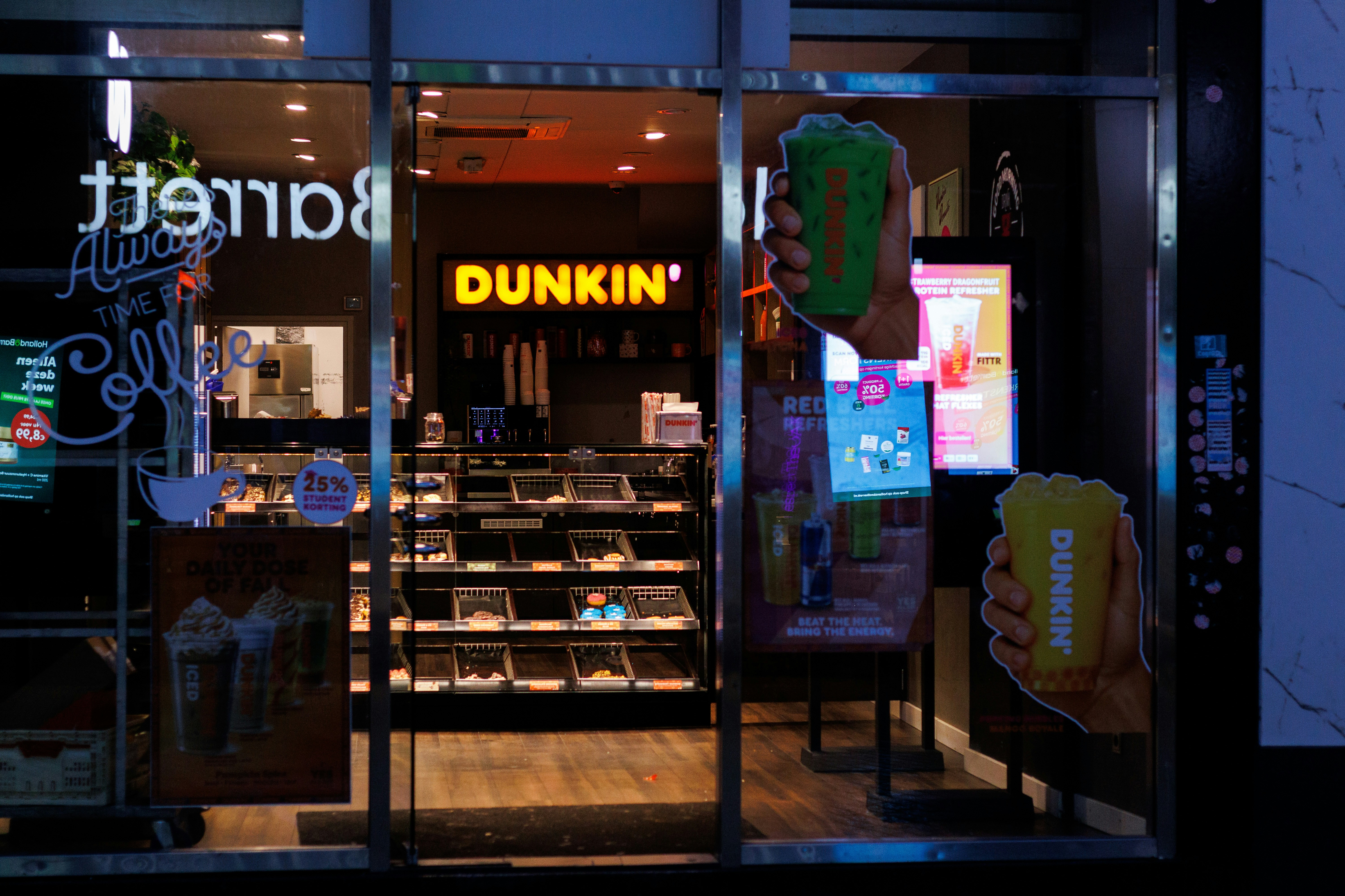 A Dunkin' shop interior viewed through a glass storefront with promotional beverage decals