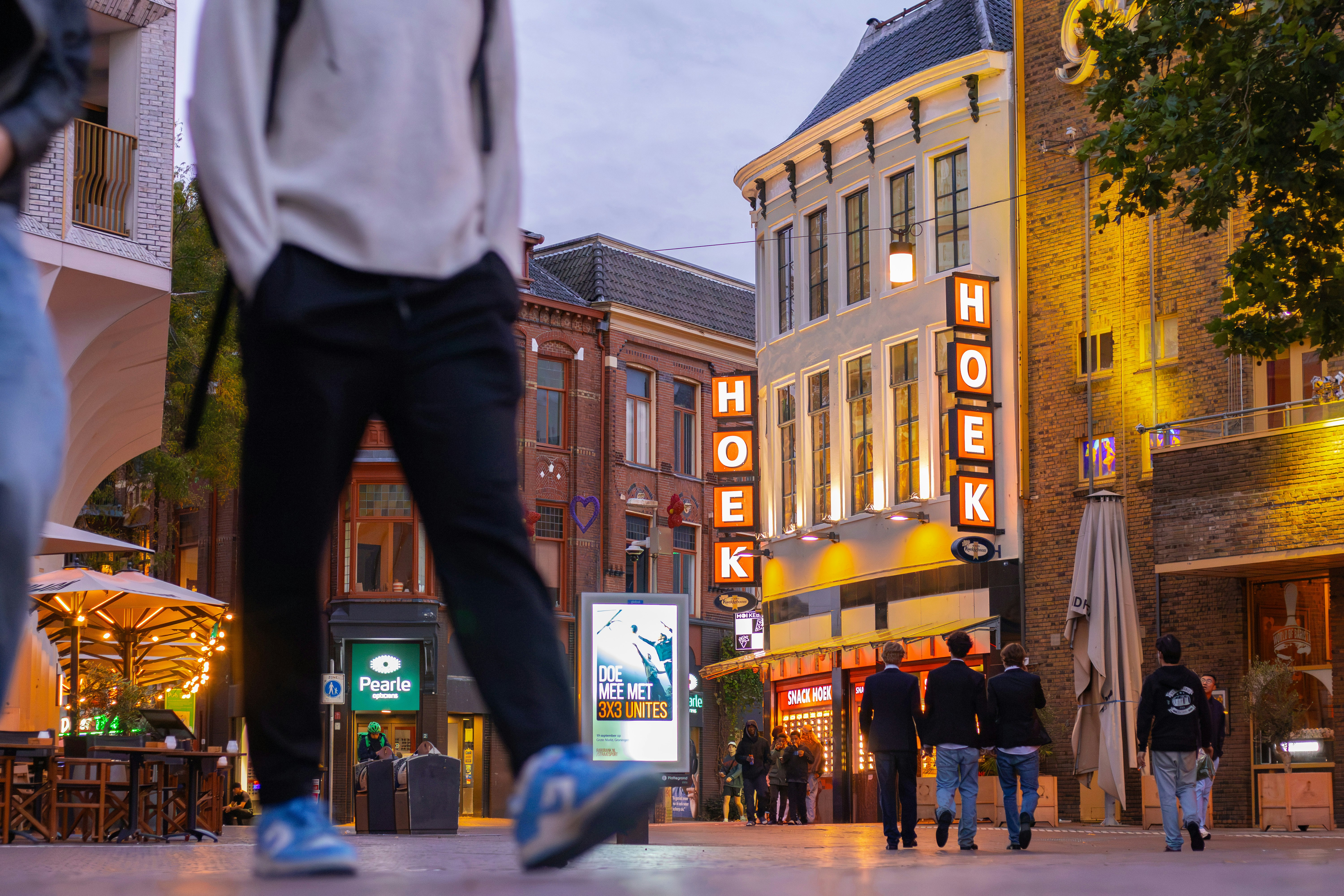 People walking on a city street with illuminated signs reading HOEK at dusk
