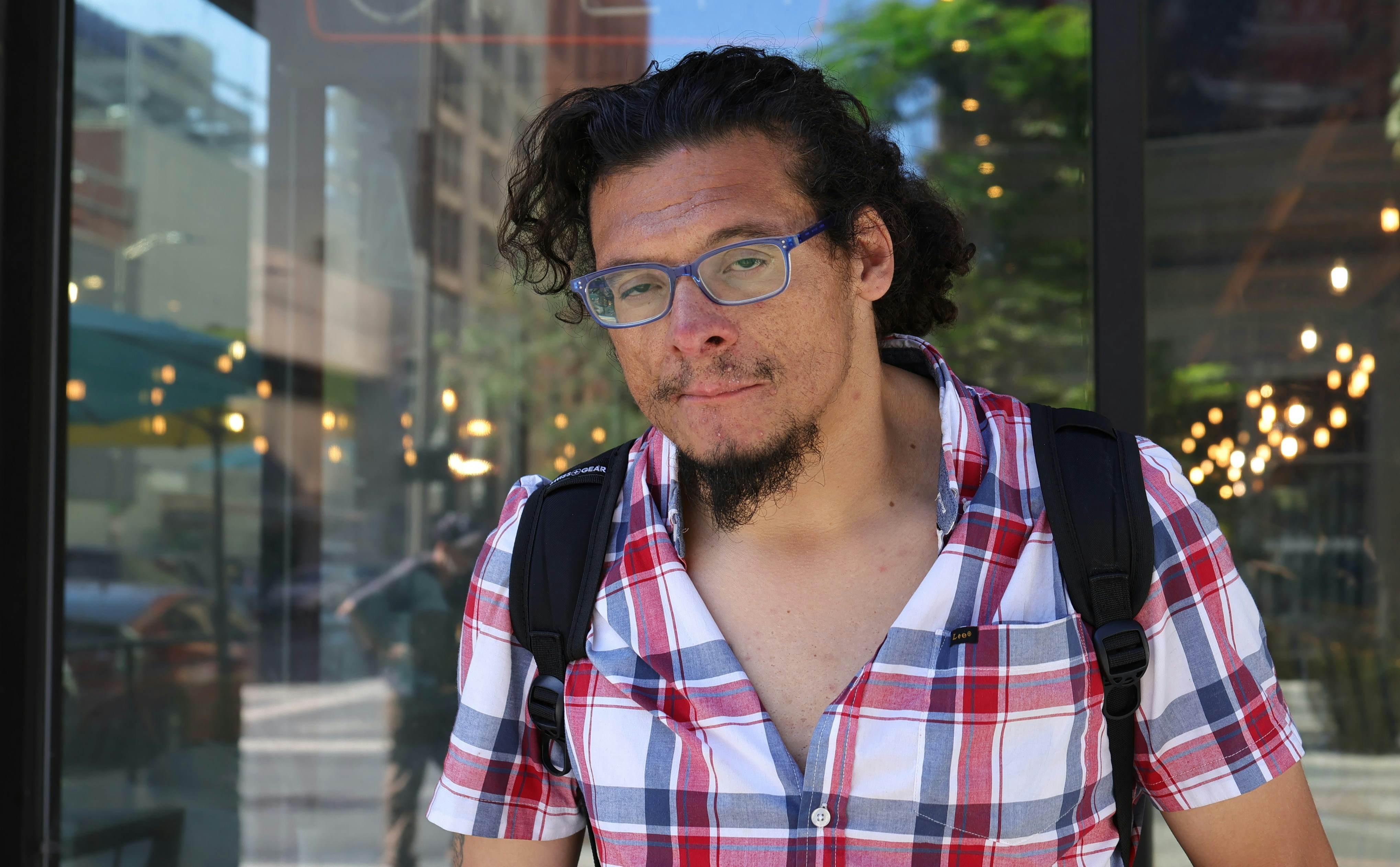 A man with a goatee and blue glasses wearing a plaid shirt and backpack