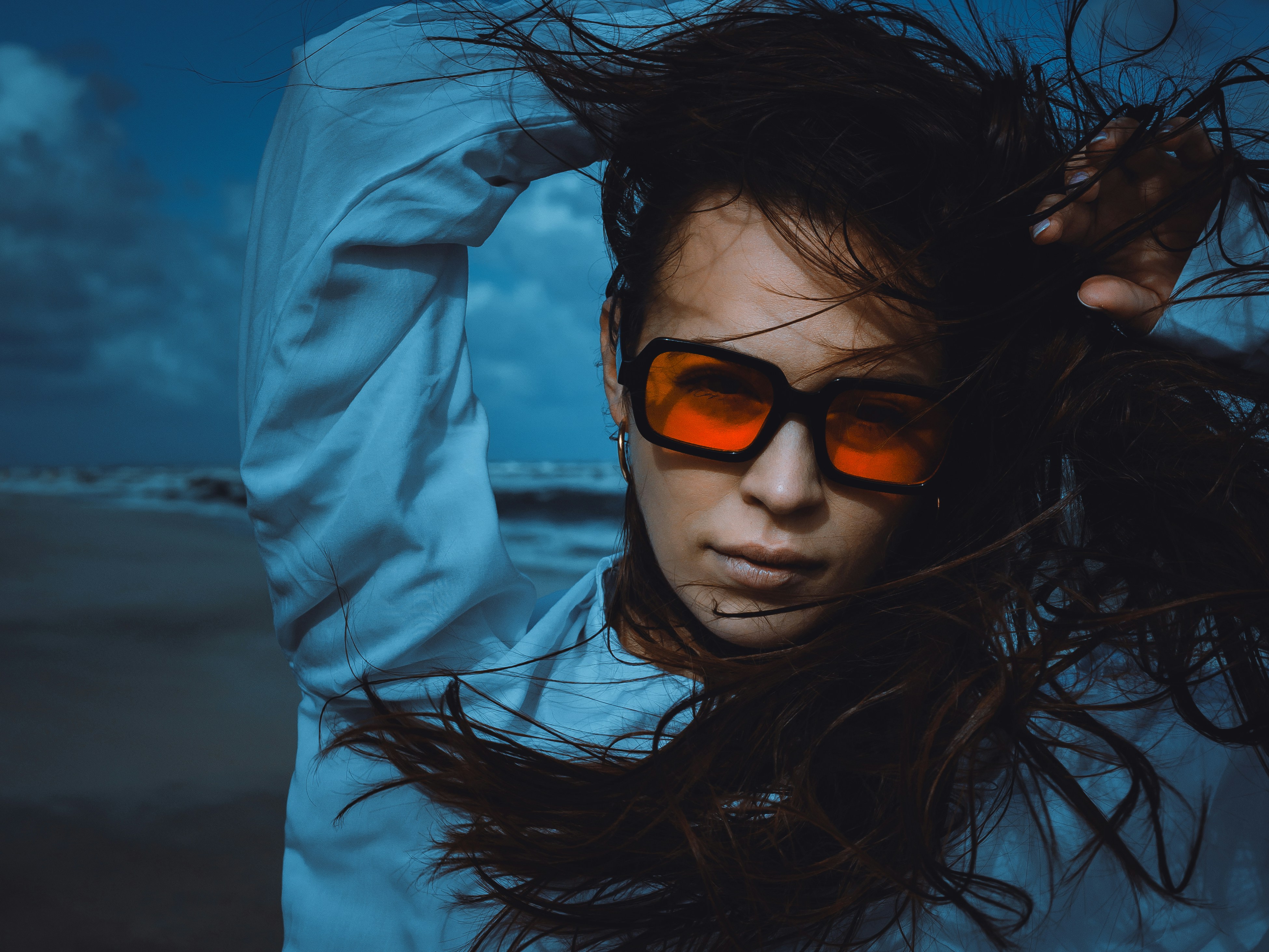 A woman with windblown hair wearing orange-tinted sunglasses on a beach at night