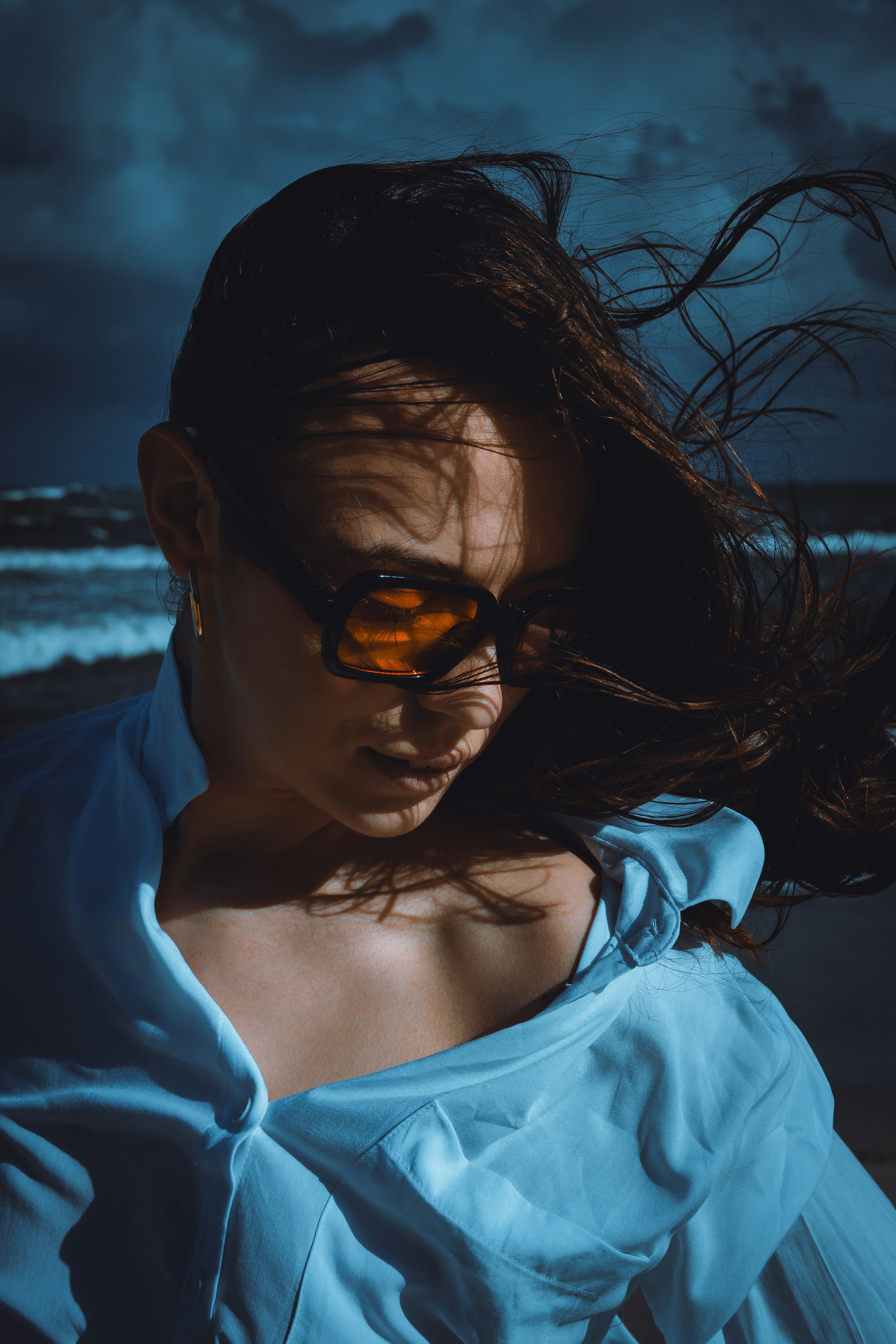 A woman with windblown hair wearing orange-tinted sunglasses and a white shirt by the sea