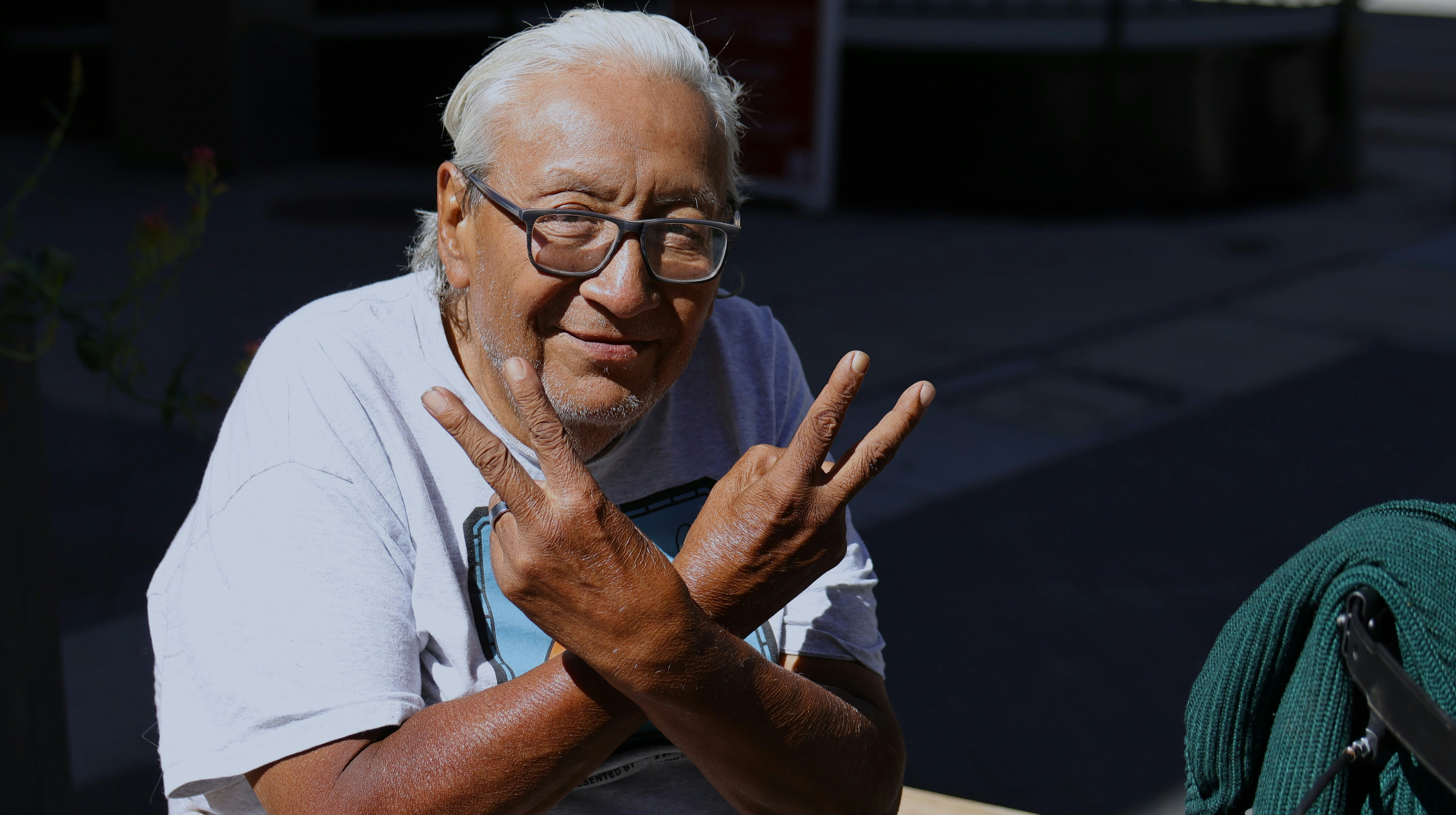 An older man with grey hair and glasses making a peace sign with both hands