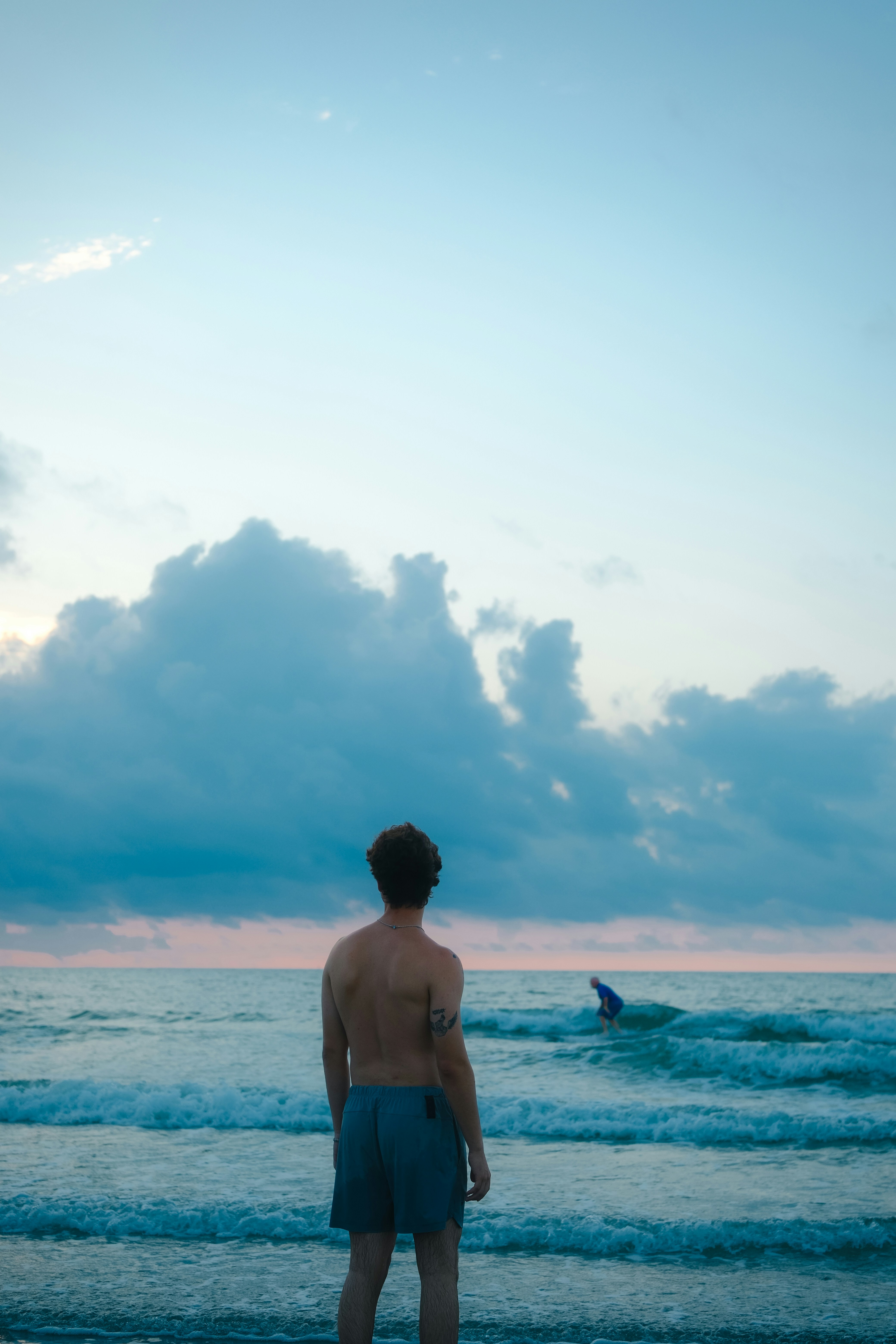 A contemplative vertical perspective captures a person standing at the shoreline, gazing out across rolling waves toward another swimmer in the distance. Towering cloud formations rise above the horizon, framing an atmospheric and peaceful coastal scene.