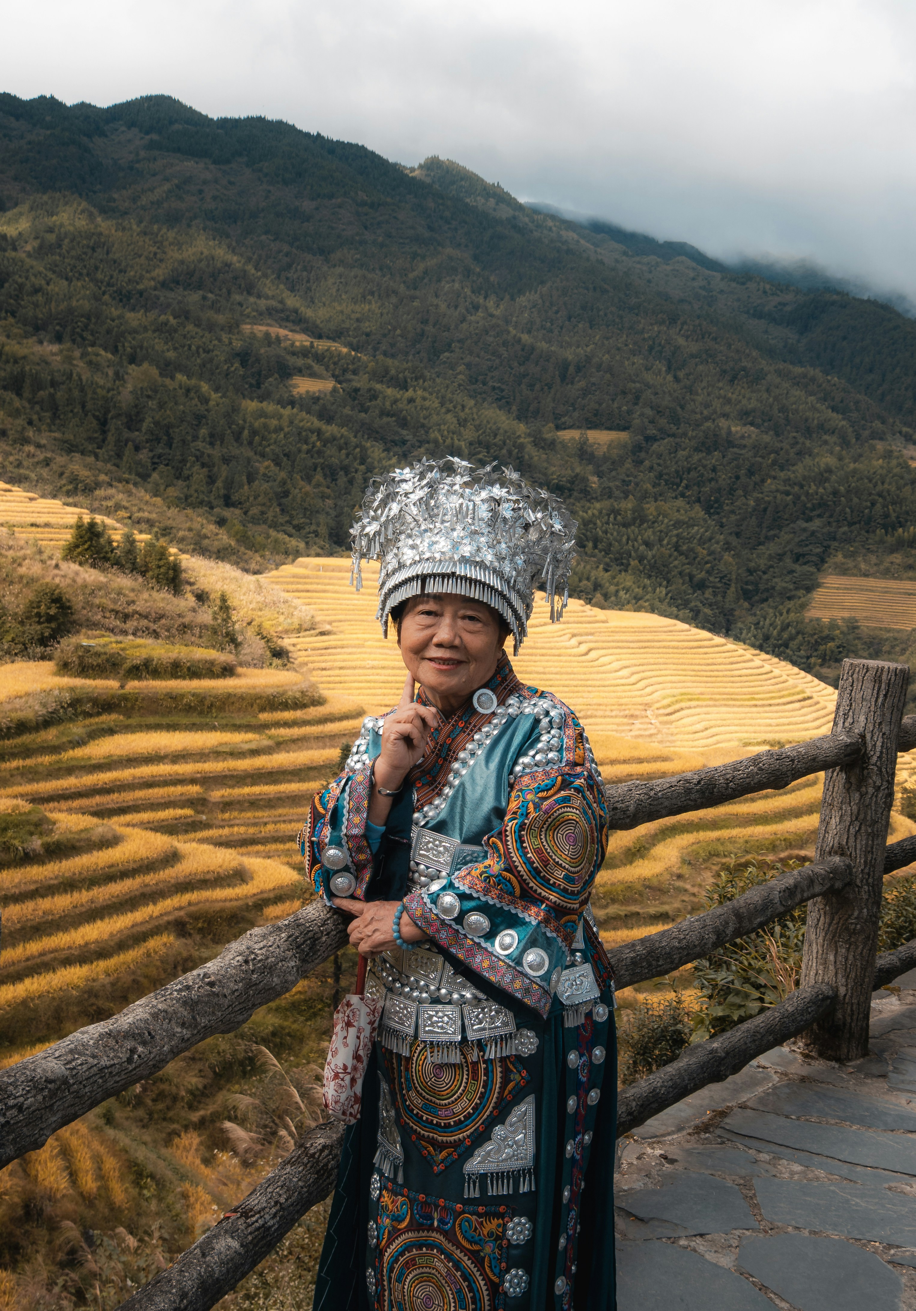 An elderly woman in traditional silver headdress and embroidered clothing by terraced rice fields