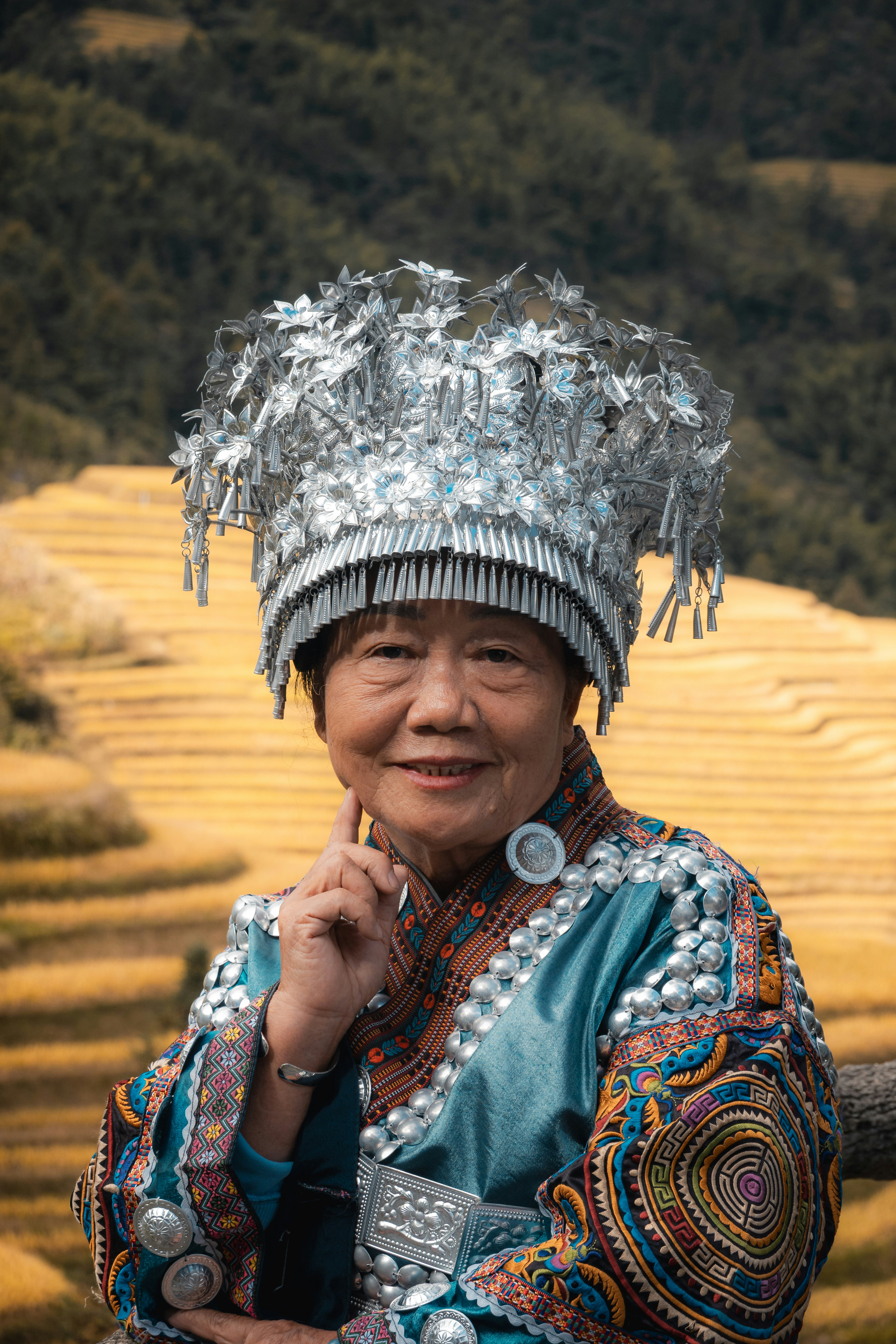 An older woman wearing a traditional silver headdress and embroidered blue clothing before rice terraces