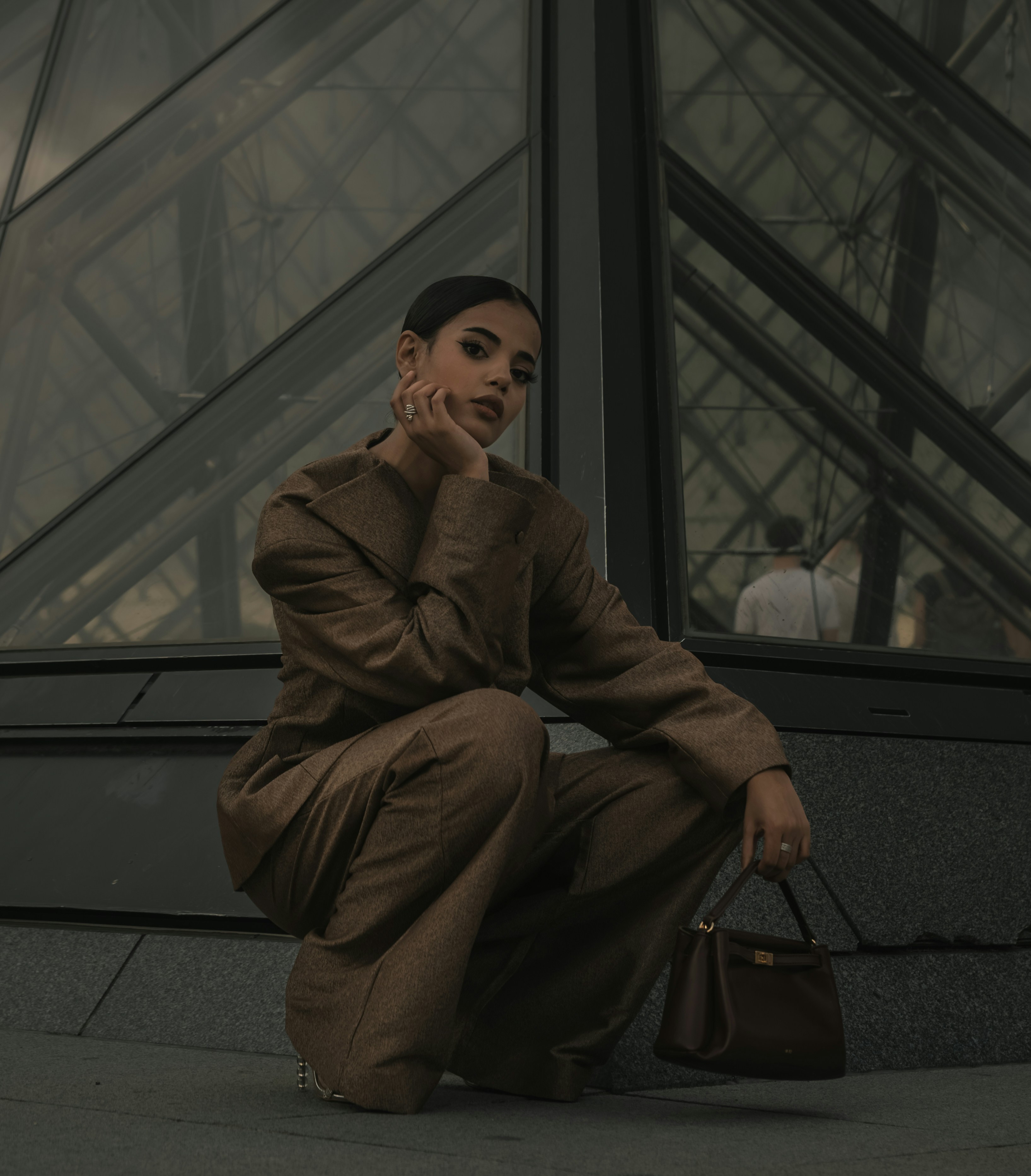 A woman in a brown suit crouching before the glass Louvre Pyramid in Paris