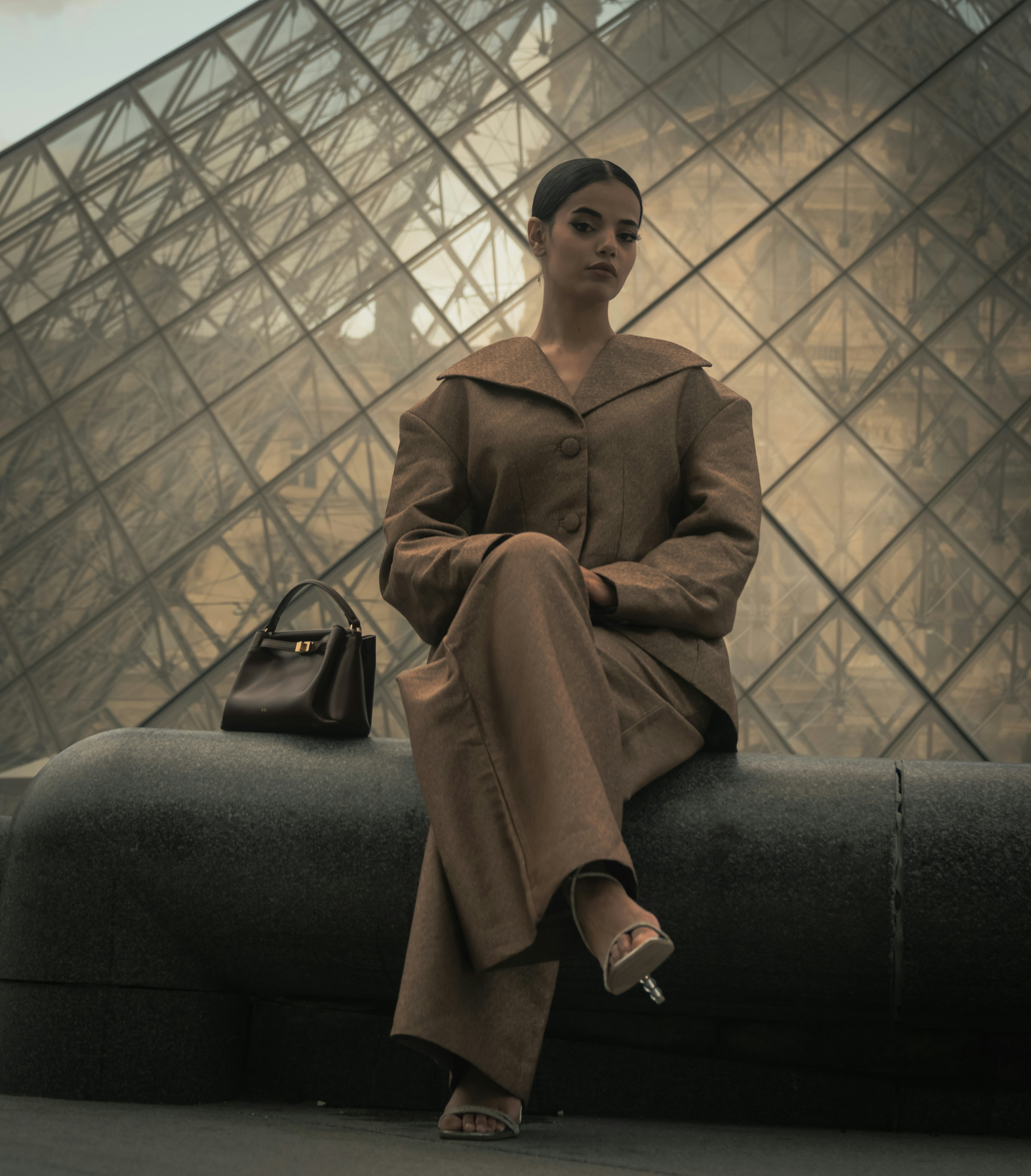 A woman in a brown suit sitting before the Louvre Pyramid in Paris