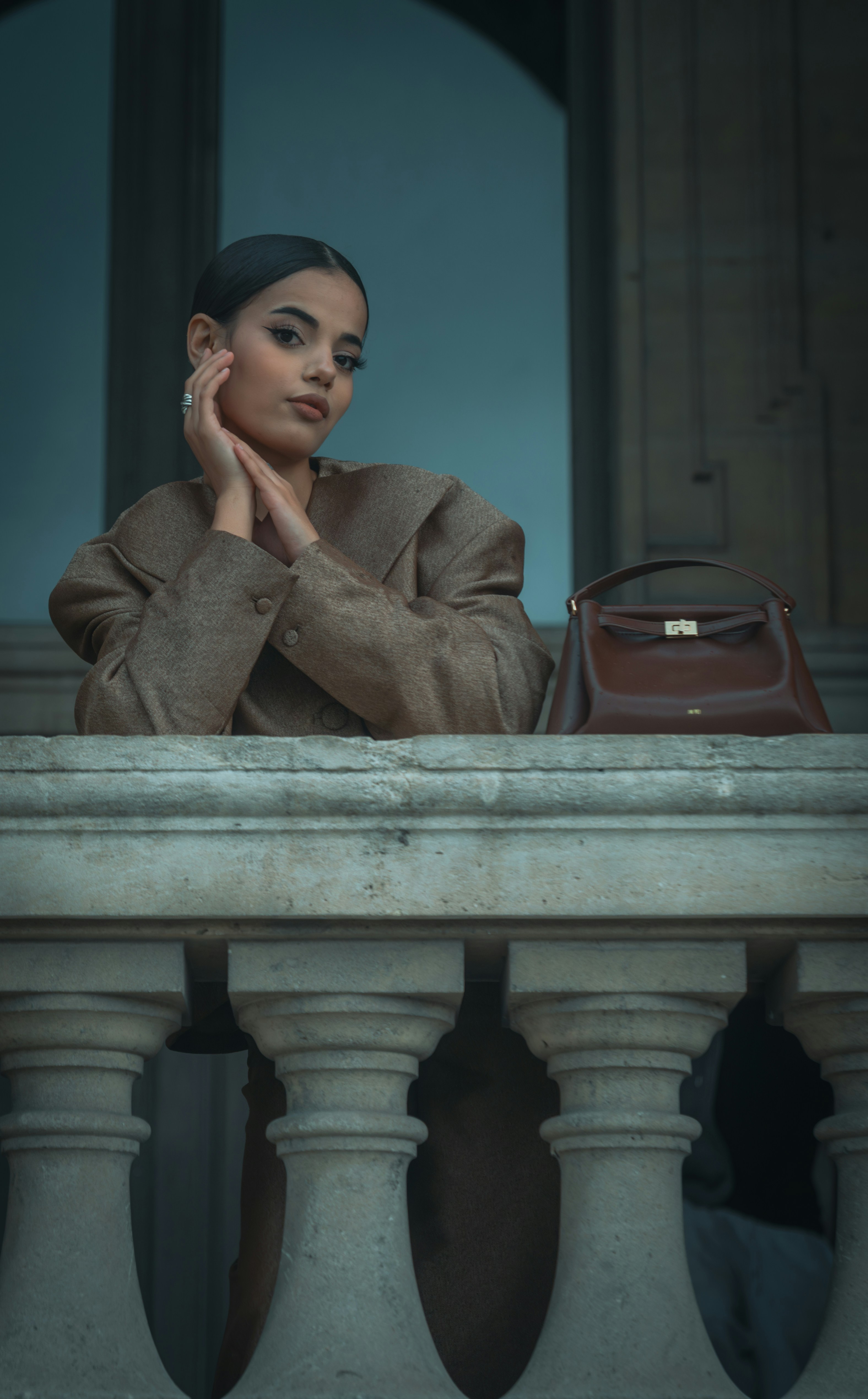 A woman in a brown blazer leaning on a stone balustrade with a handbag